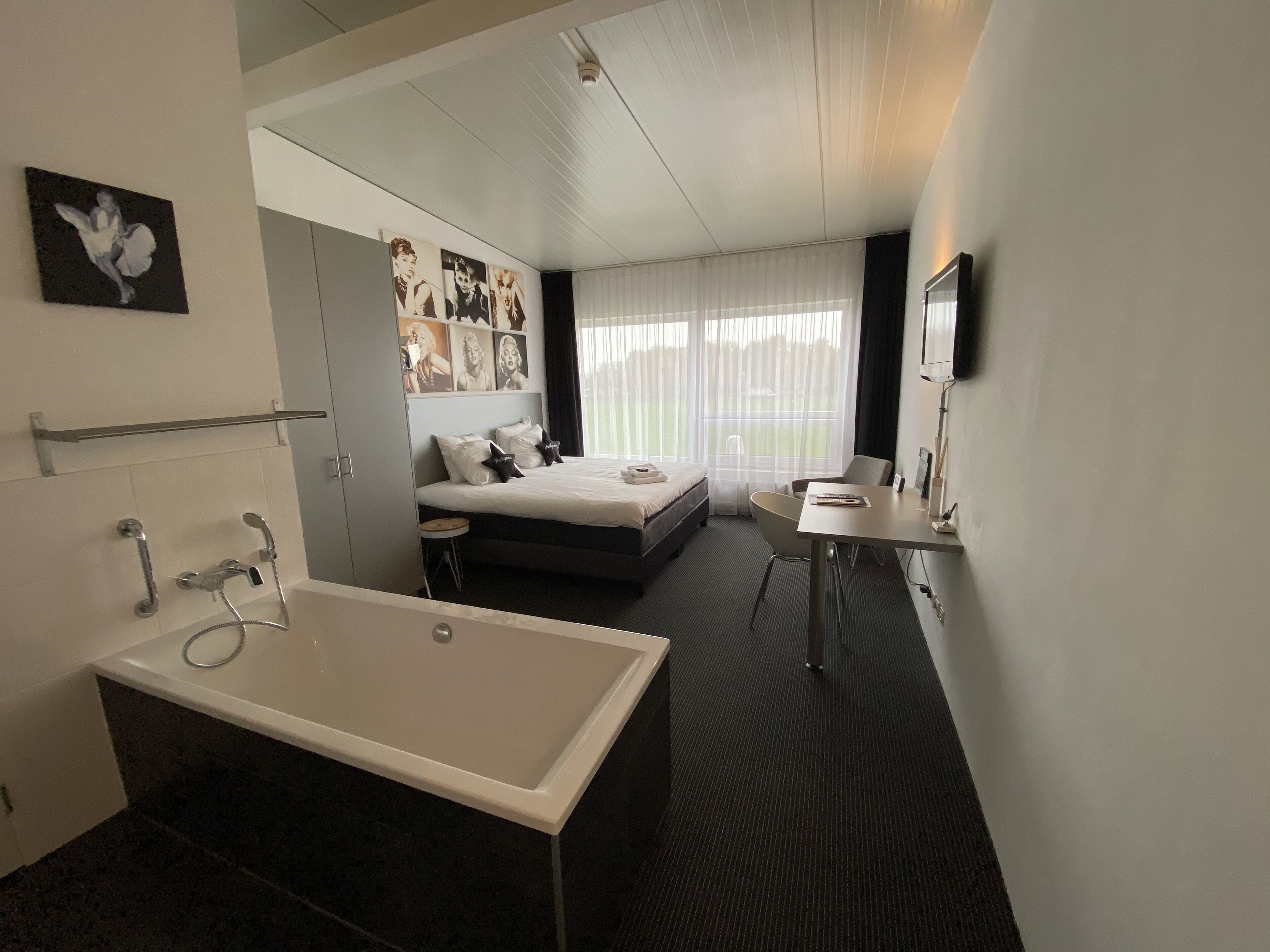 A modern hotel room with a large bathtub, a bed with two pillows, a desk with a chair, and a large window with sheer curtains.