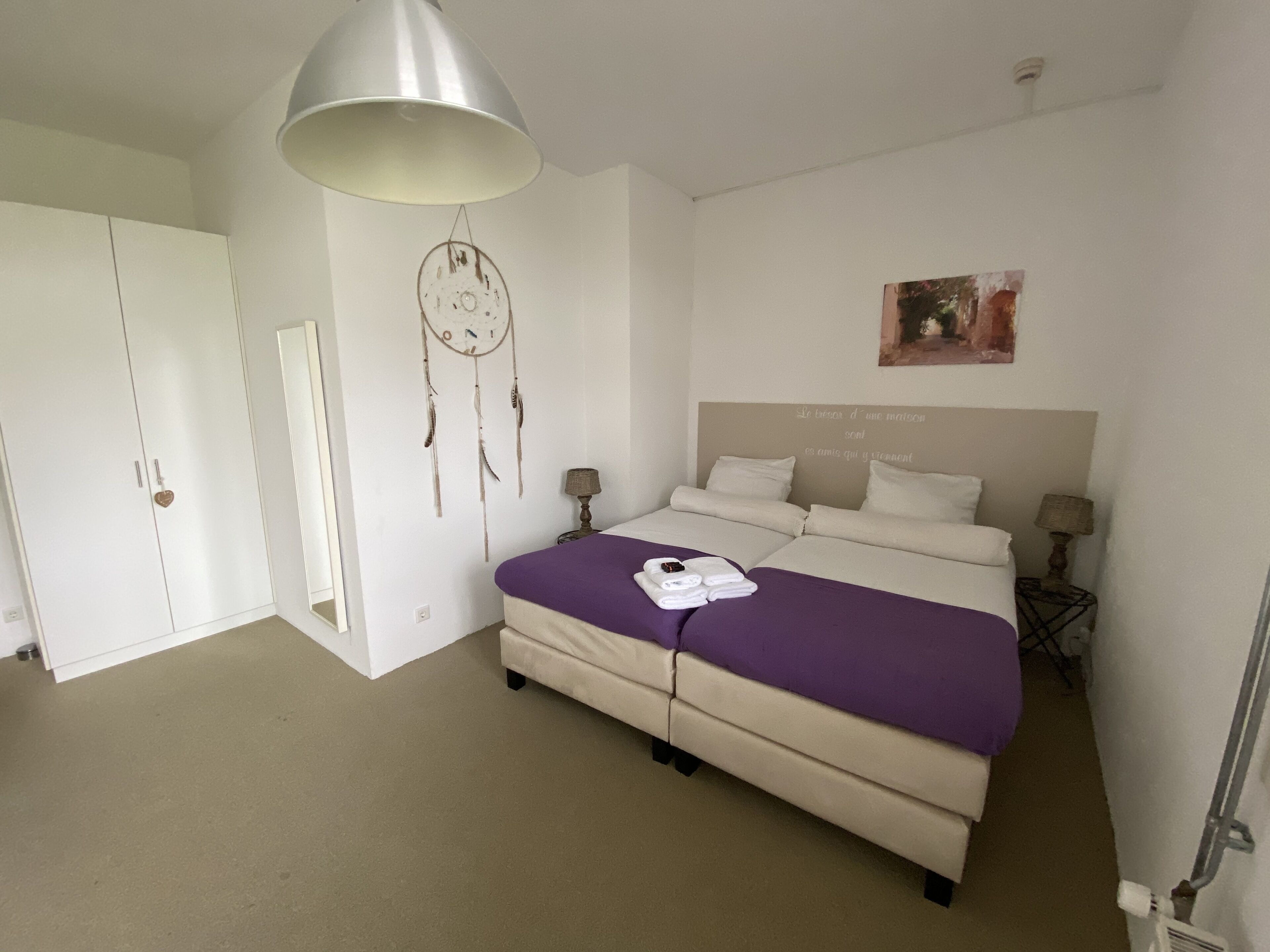 Une chambre à coucher avec un lit, des tables de chevet, une armoire, un attrape-rêves et un mur orné d’un message.