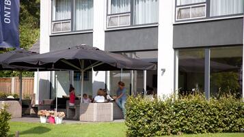 Un área de estar al aire libre con mobiliario de mimbre y sombrillas, un edificio con amplios ventanales y un seto en primer plano.