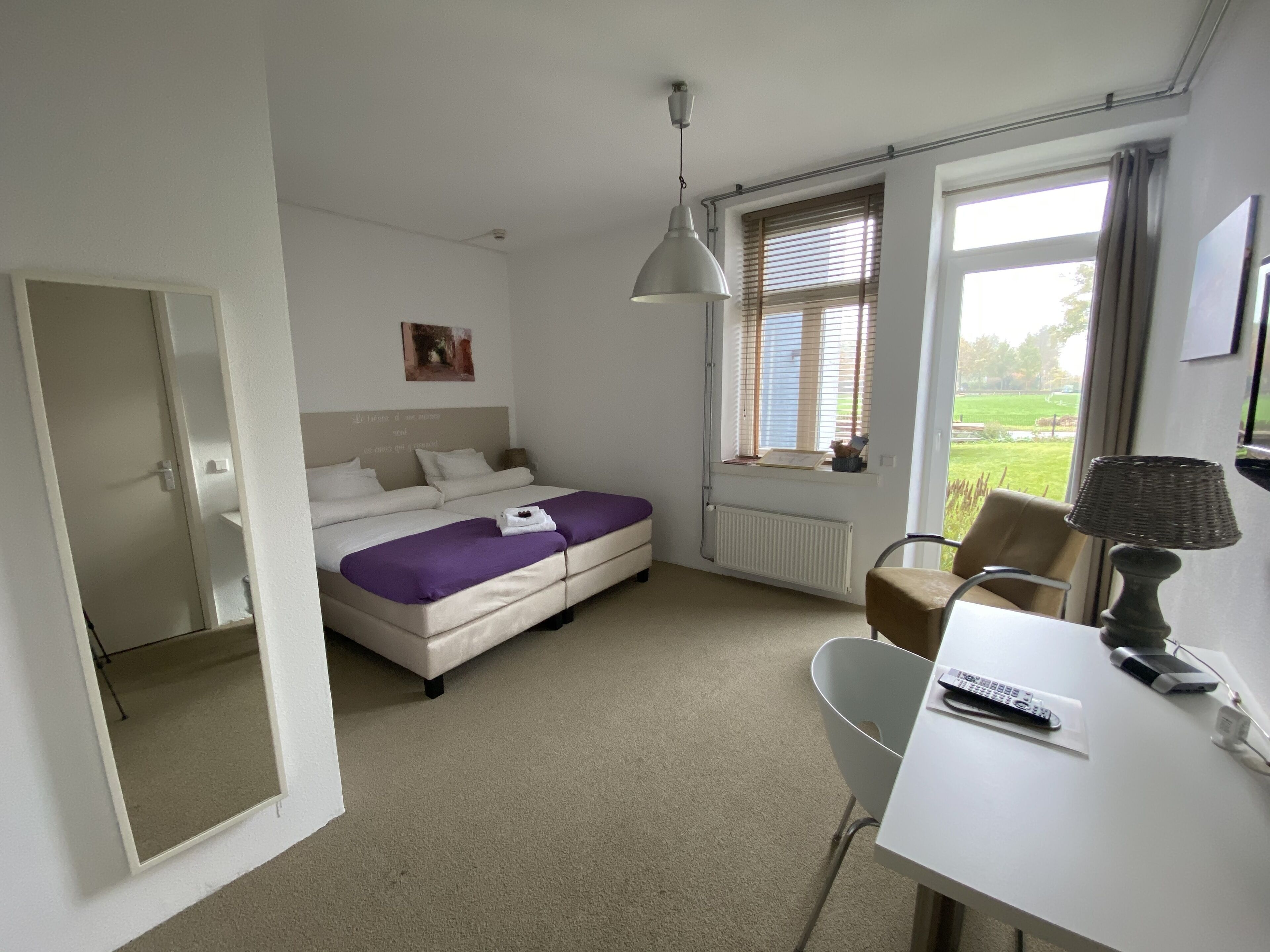 Ein Schlafzimmer mit einem Bett, einem Schreibtisch, einem Stuhl, einer Lampe, einem Fenster mit Jalousien und Blick ins Freie.