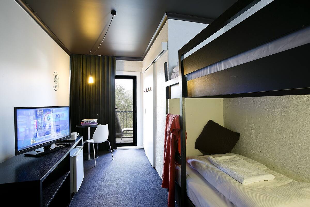 A hotel room with a bunk bed, a desk with a television, a chair, and a small table with books.