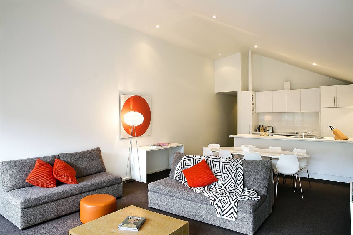 A modern living room with a grey sofa, orange cushions, and a coffee table. A dining area with white chairs and a kitchen in the background.