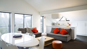 A modern living room with a white table, grey sofas with orange cushions, and a kitchen area with white cabinets and a sink. - Silverski (Falls Creek)