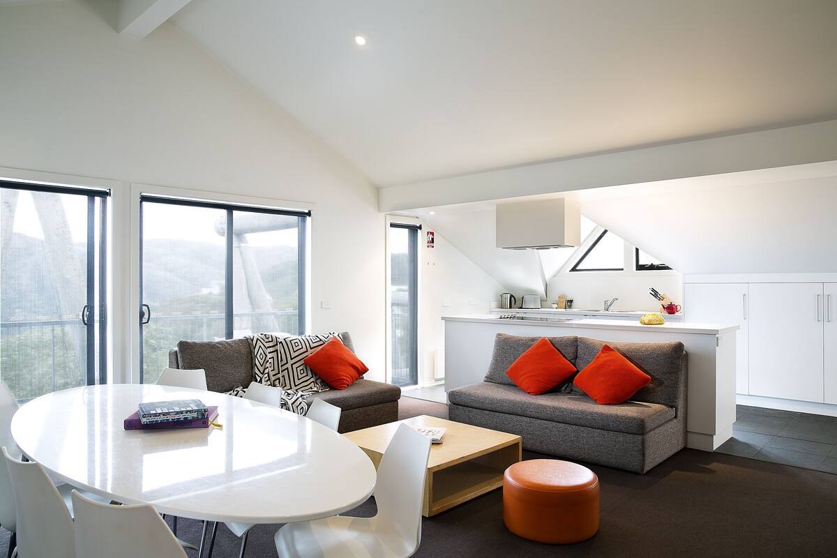 A modern living room with a white table, grey sofas with orange cushions, and a kitchen area with white cabinets and a sink.