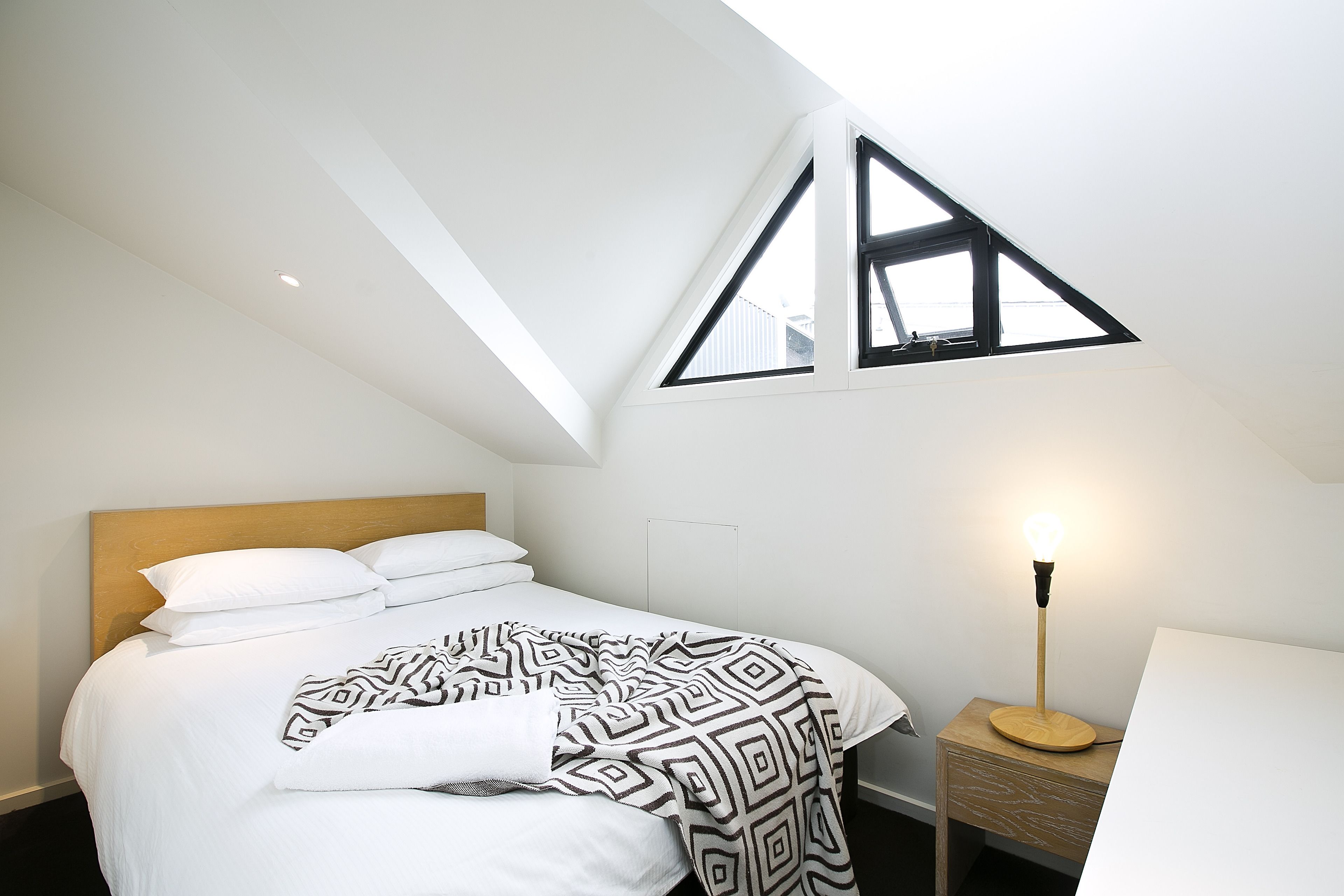 A bedroom with a sloped ceiling, a bed with white bedding, a patterned throw blanket, a bedside lamp, and a small wooden nightstand.