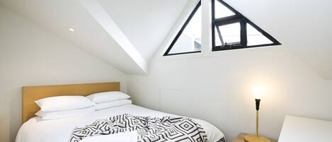 A bedroom with a sloped ceiling, a bed with white bedding, a patterned throw blanket, a bedside lamp, and a small wooden nightstand.