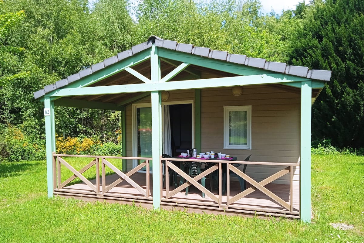 Uma pequena cabana de madeira com varanda, uma mesa e cadeiras, e uma janela.