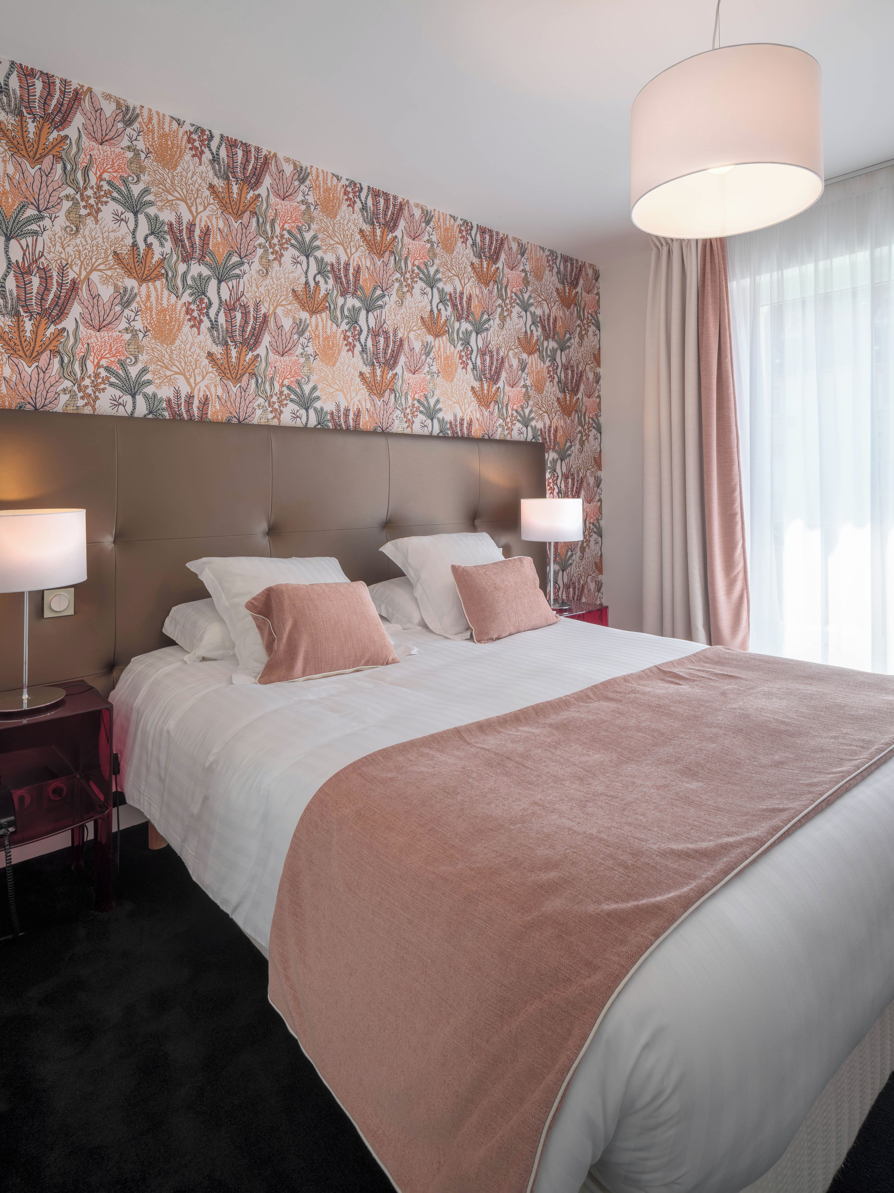 Een hotelkamer met een groot bed, twee nachtkastjes, een wand met bloemenmotief en een groot raam met gordijnen.