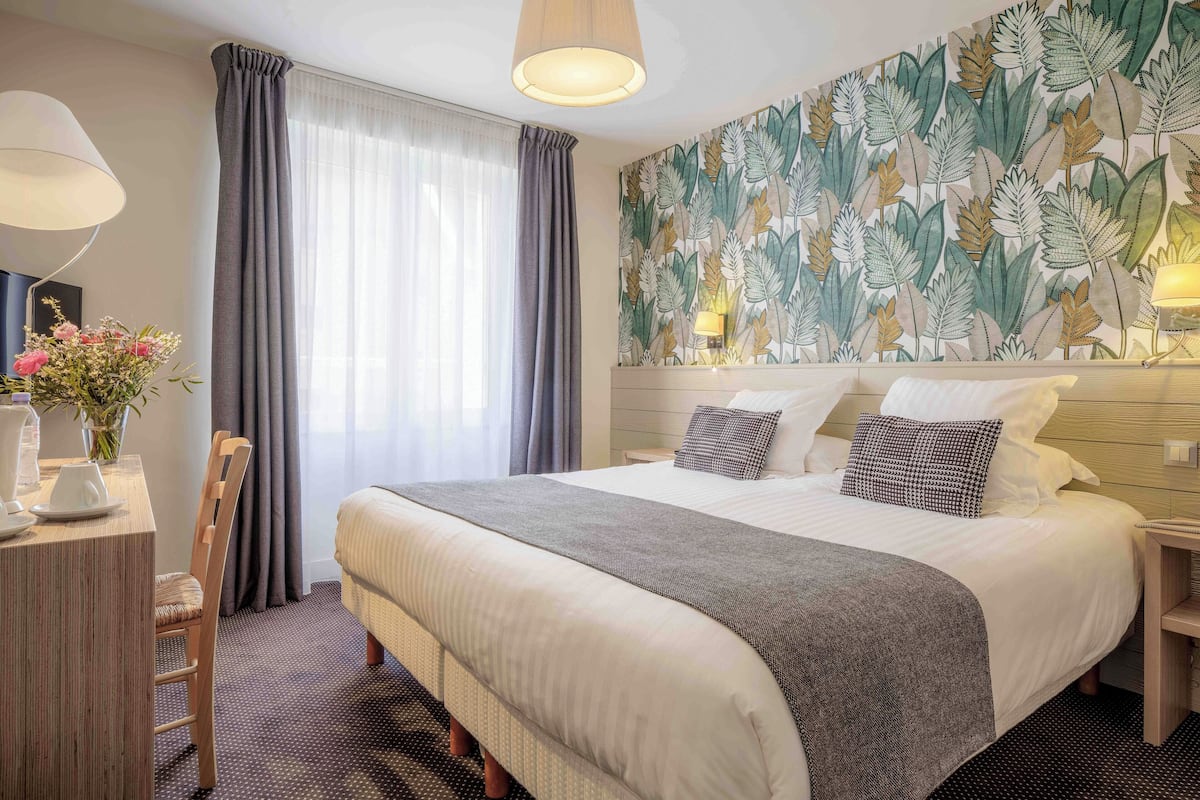 A hotel room with a large bed, a desk with a chair, a vase of flowers, and a wall with a leaf pattern.