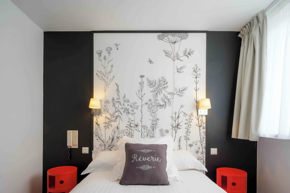 A hotel room with a large bed, two bedside lamps, a red nightstand, and a floral wallpaper.