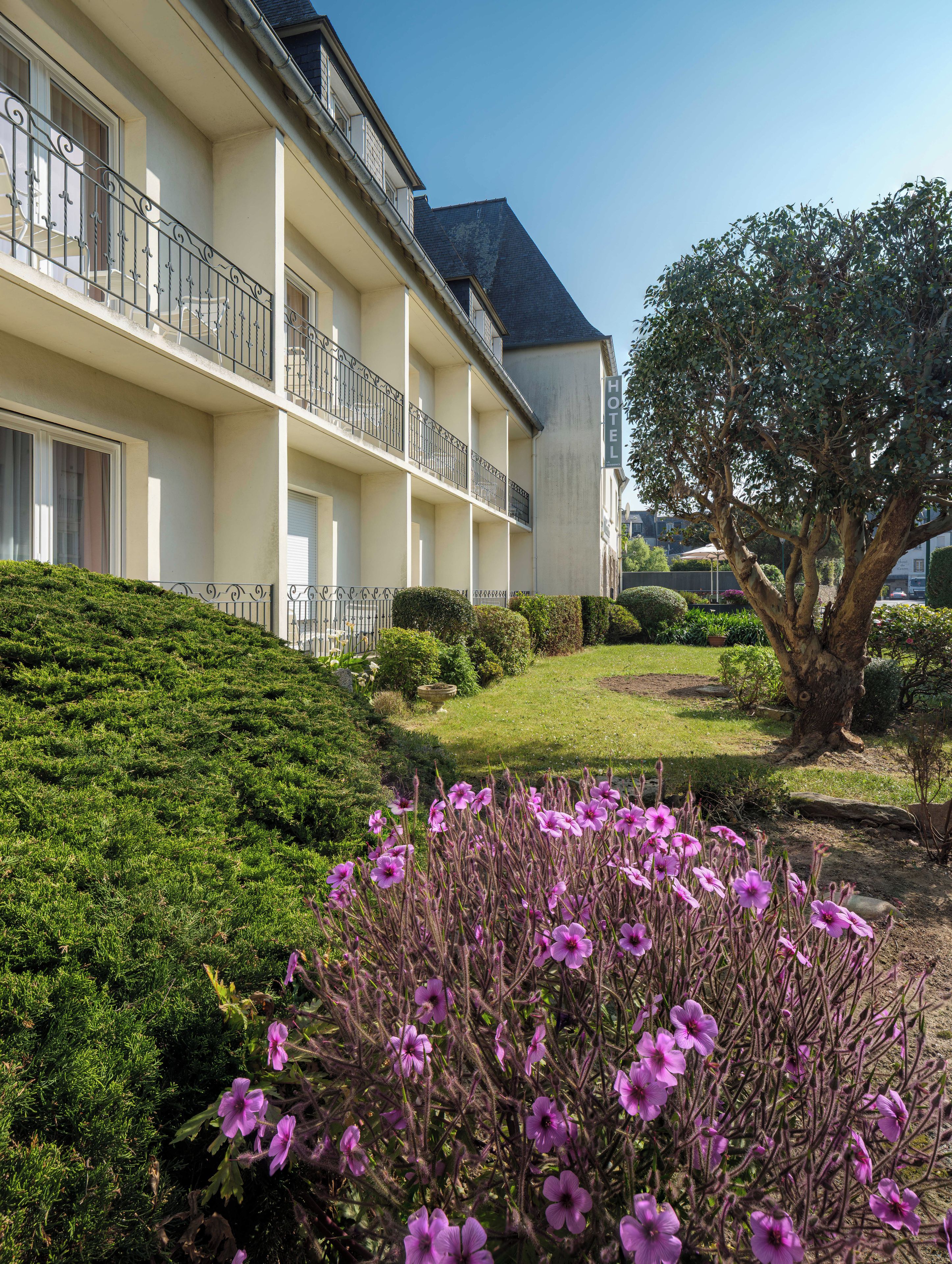 Un hotel con varios balcones, un jardín cuidado con arbustos y flores, y un árbol grande.