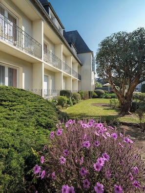 A hotel with multiple balconies, a well-maintained garden with shrubs and flowers, and a large tree.