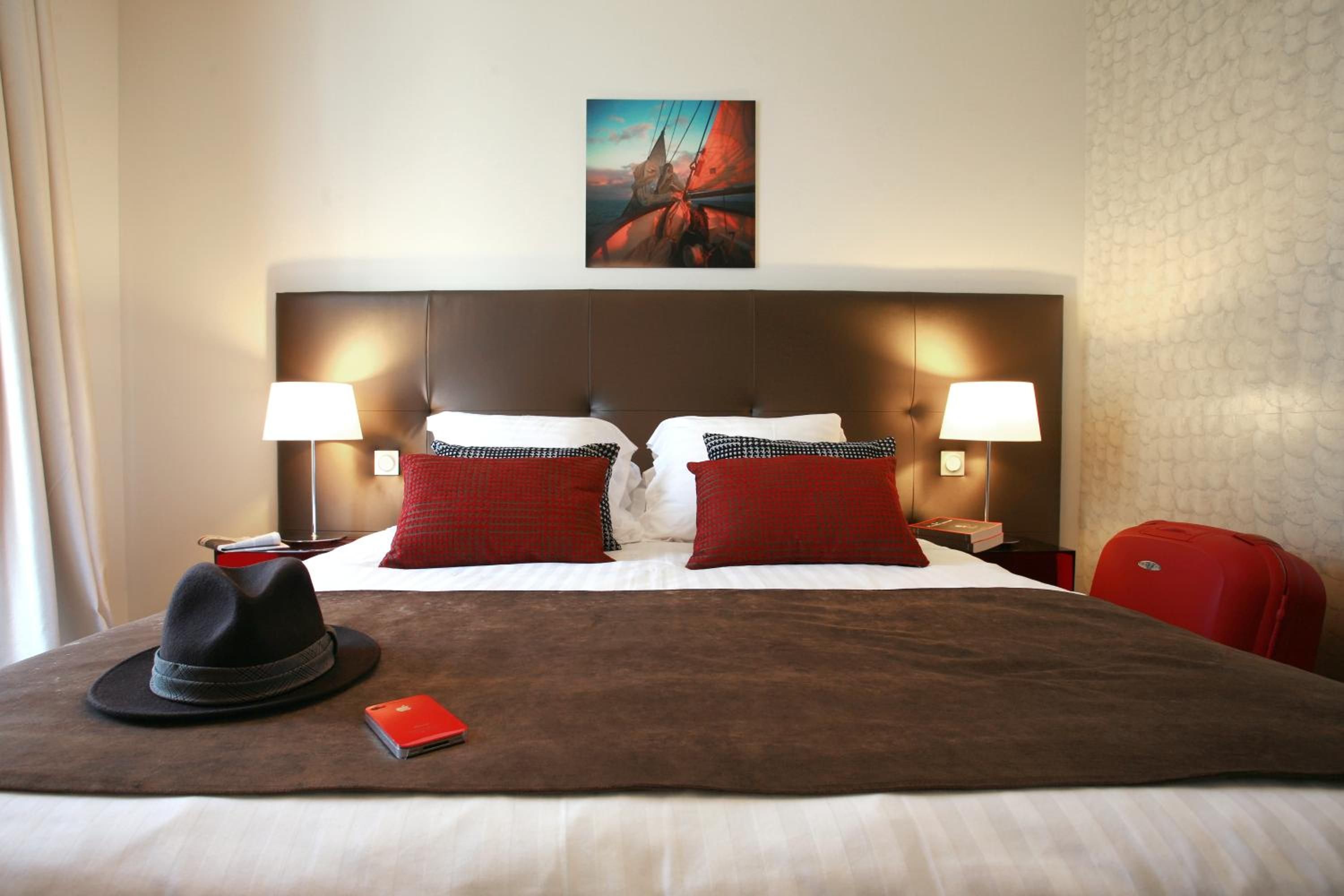 A hotel room with a large bed, two bedside lamps, a painting above the bed, and a red suitcase on the floor.
