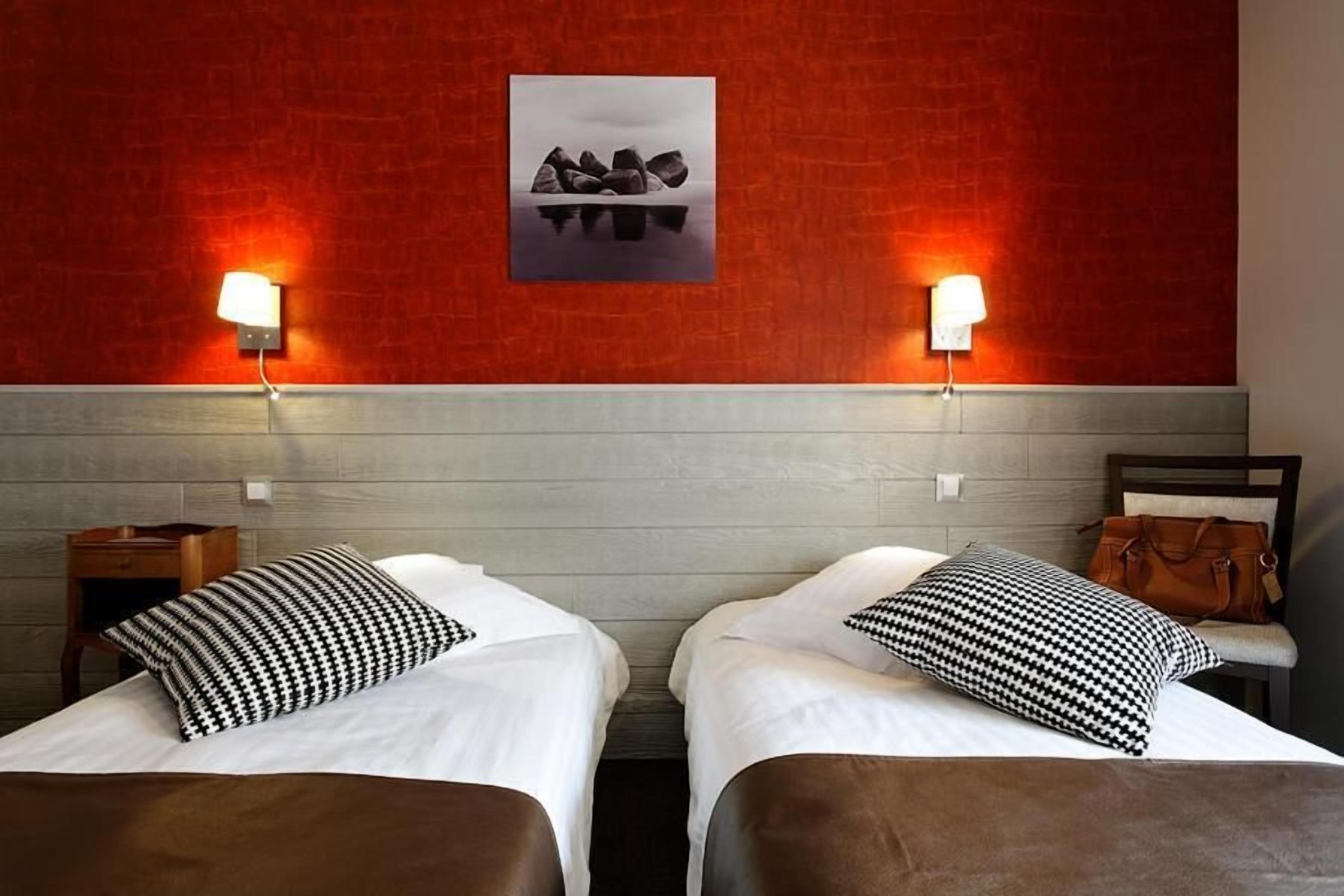 A hotel room with two beds, each with a white bedspread and black-and-white patterned pillows. There are two wall-mounted lamps on either side of a framed picture.