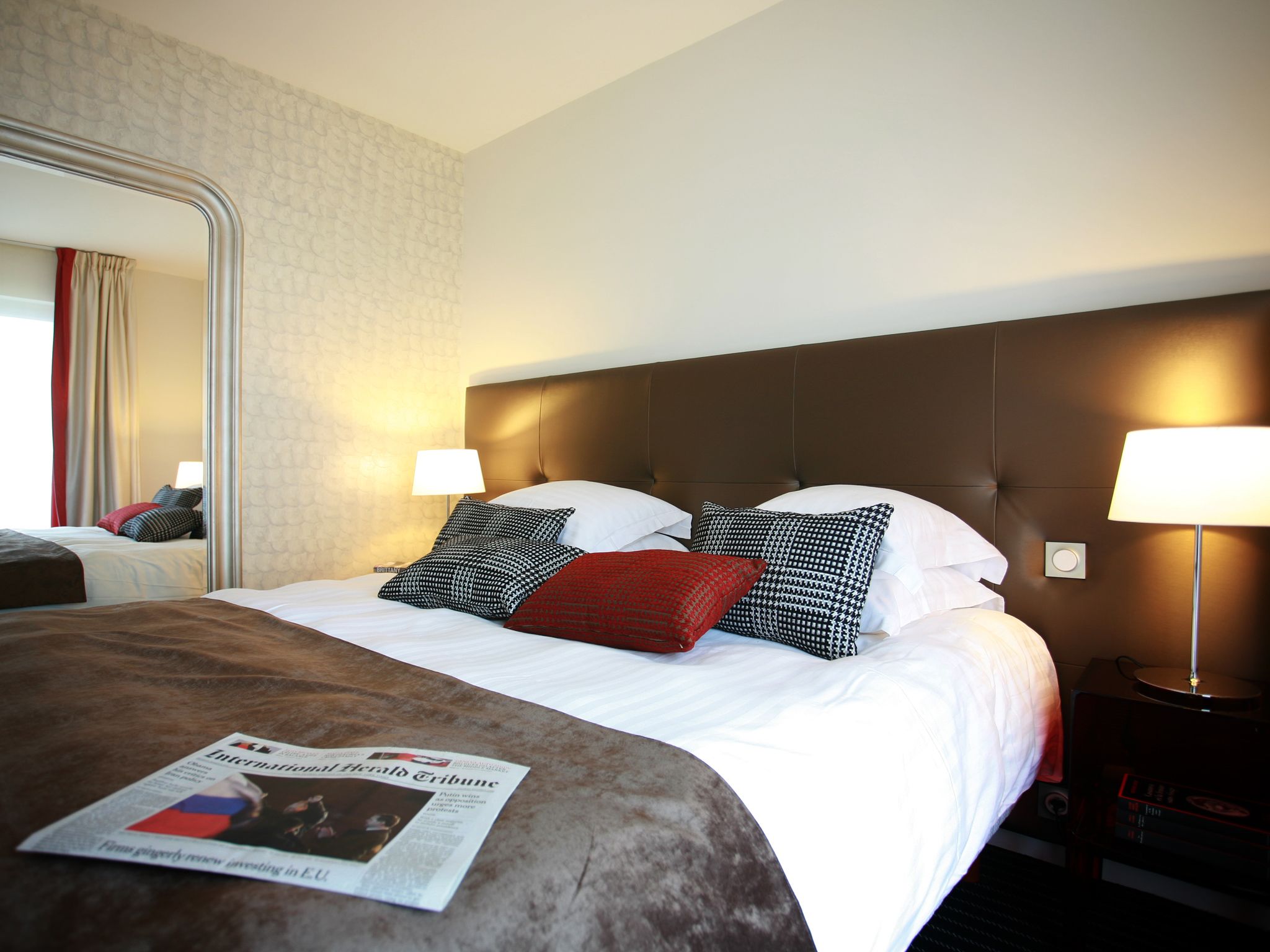 A hotel room with a large bed, two bedside lamps, a mirror, and a newspaper on the bed.