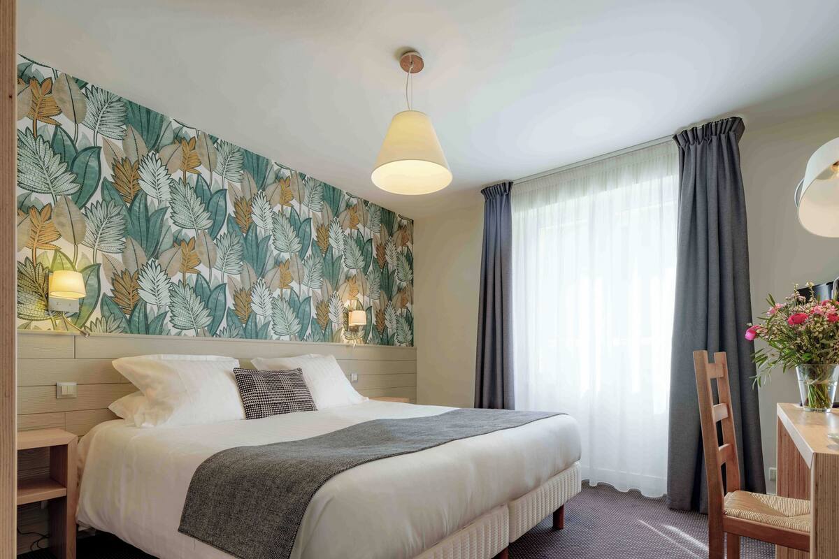 A neatly made bed with a grey blanket, a wall with a leafy pattern, a bedside lamp, and a chair with a vase of flowers.