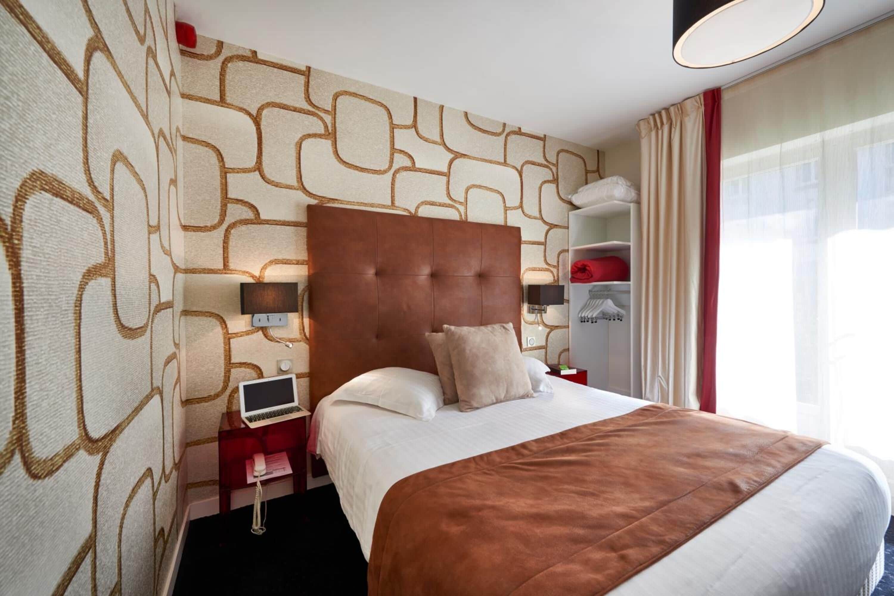 Habitación de hotel con una cama grande, cabecero, mesita de noche con lámpara, armario y ventana con cortinas.