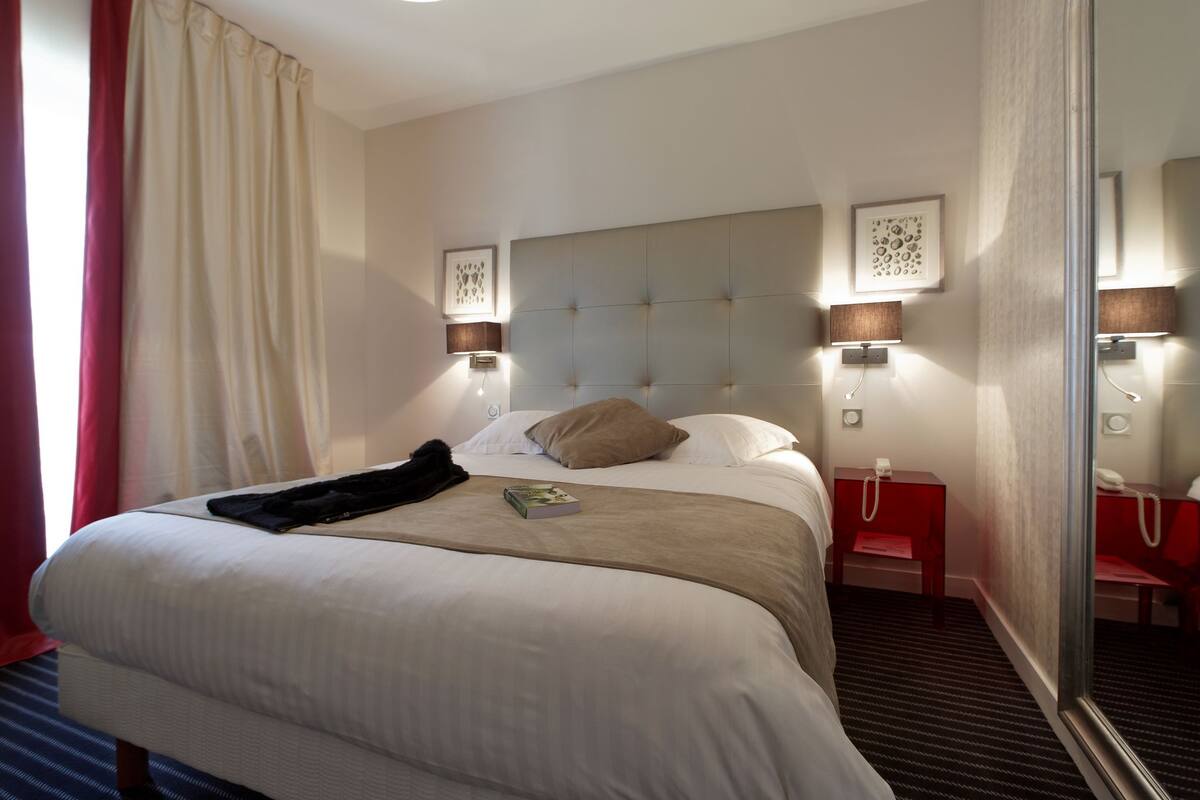 A hotel room with a large bed, two bedside tables, a red nightstand, a mirror, and a window with curtains.