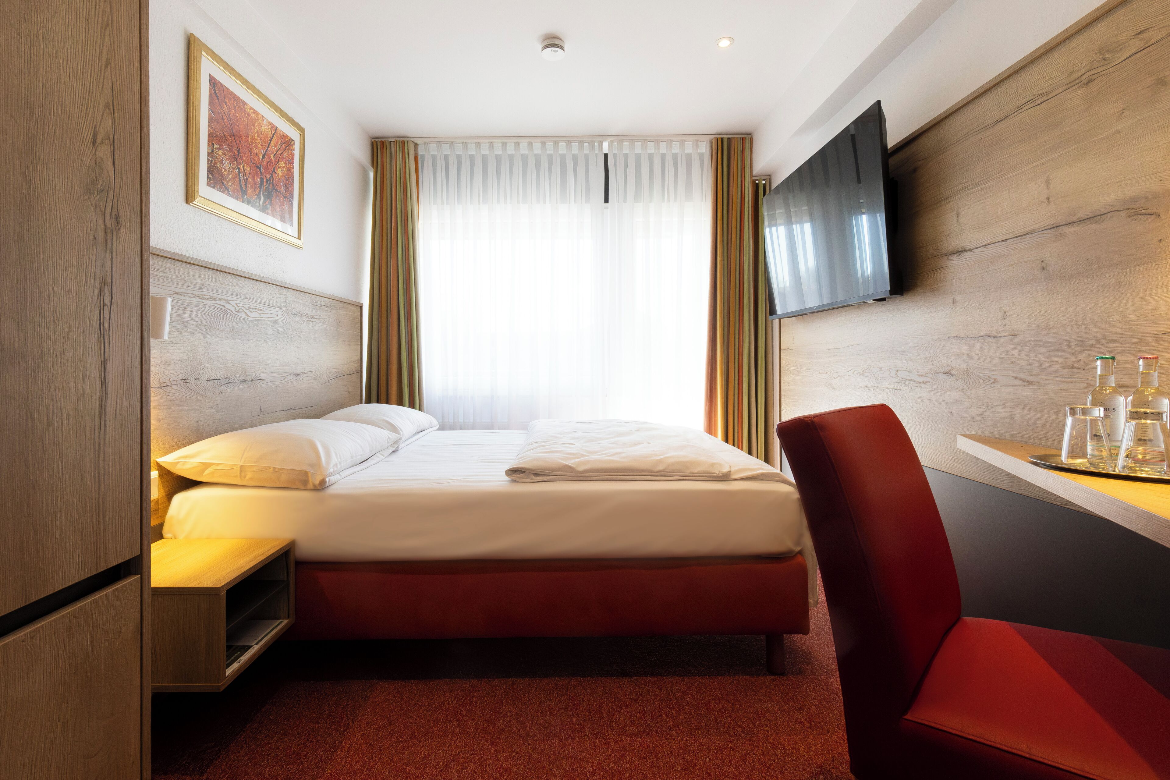 A hotel room with a bed, a TV mounted on the wall, a desk with a chair, and a window with curtains.