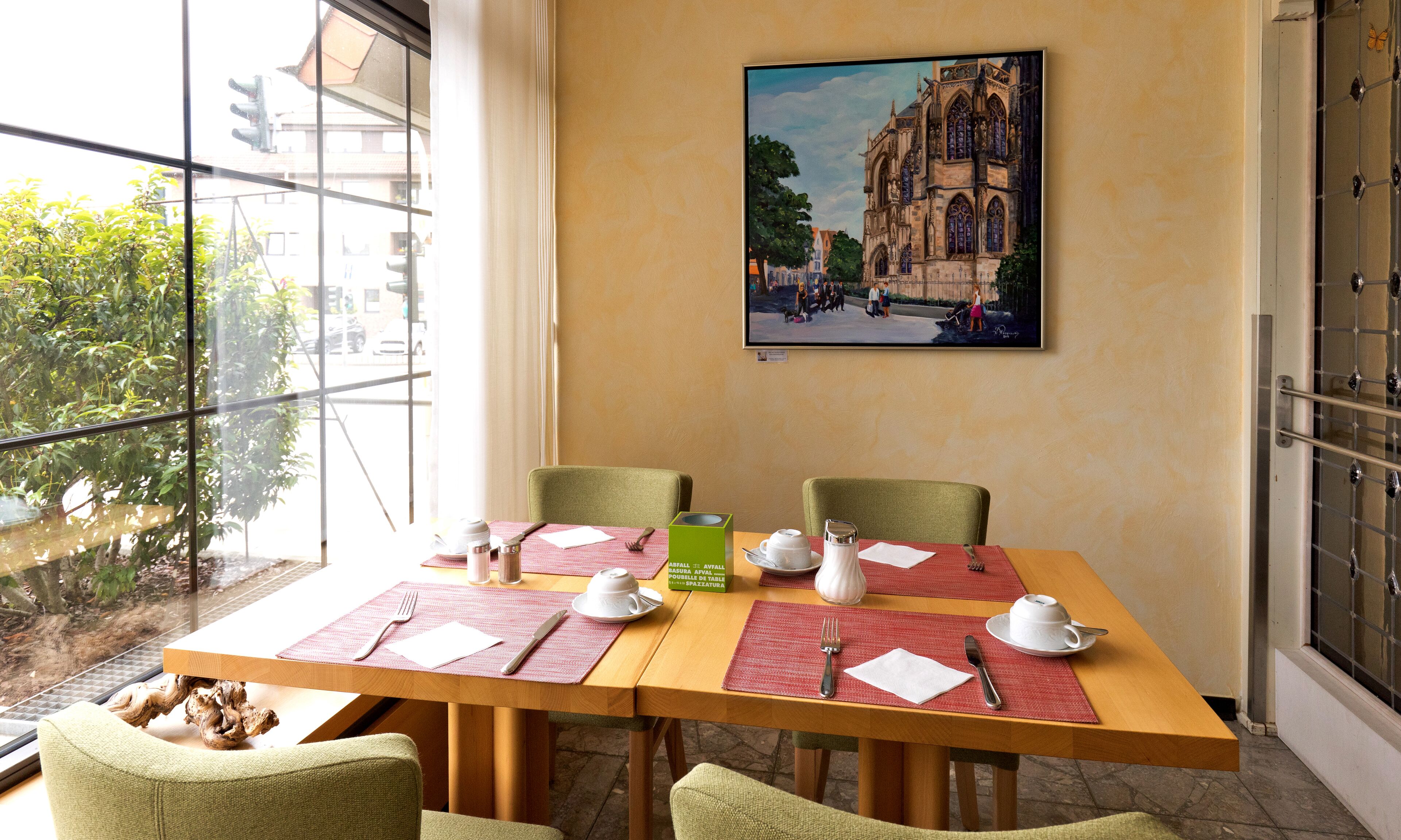 A dining table set with placemats, cutlery, and a teapot, with a painting of a building on the wall.