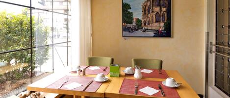 Una mesa de comedor dispuesta con manteles individuales, cubiertos y una tetera, con un cuadro de un edificio en la pared.