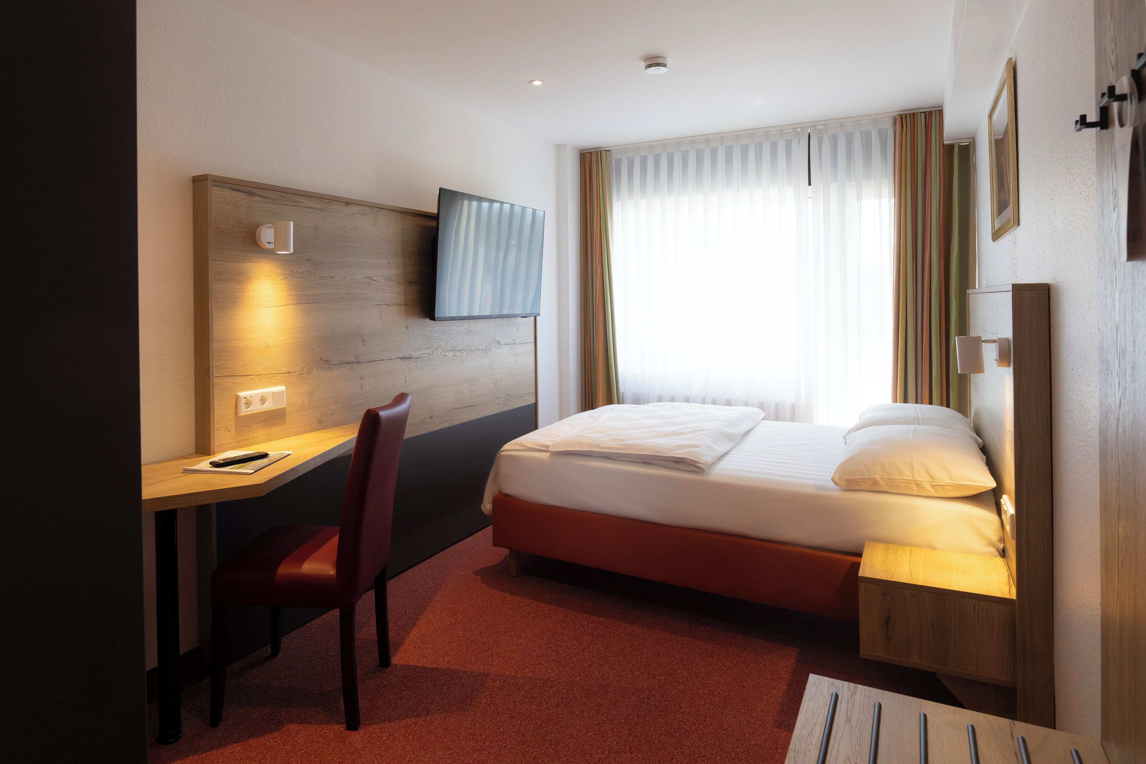 Quarto de hotel com cama, escrivaninha, cadeira, televisão e cortinas.