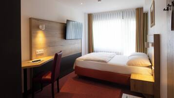 Quarto de hotel com cama, escrivaninha, cadeira, televisão e cortinas.