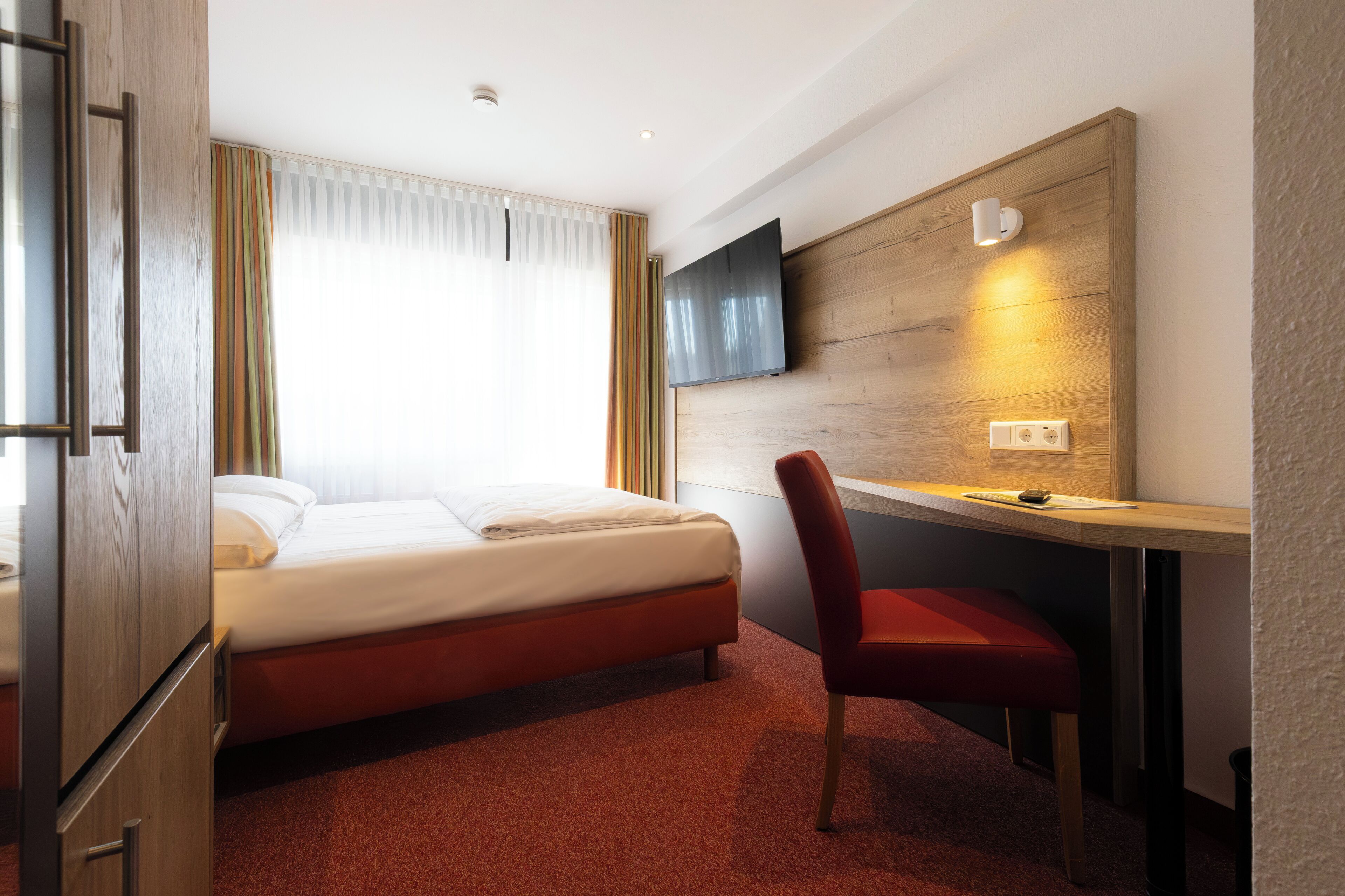 Une chambre d’hôtel équipée d’un lit, d’un bureau, d’une chaise, d’un téléviseur fixé au mur et d’une fenêtre avec des rideaux.