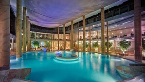 A multi-level indoor swimming pool with a central water feature, surrounded by seating areas and a glass ceiling. - Hotel Mariaweiler Hof (Dueren)