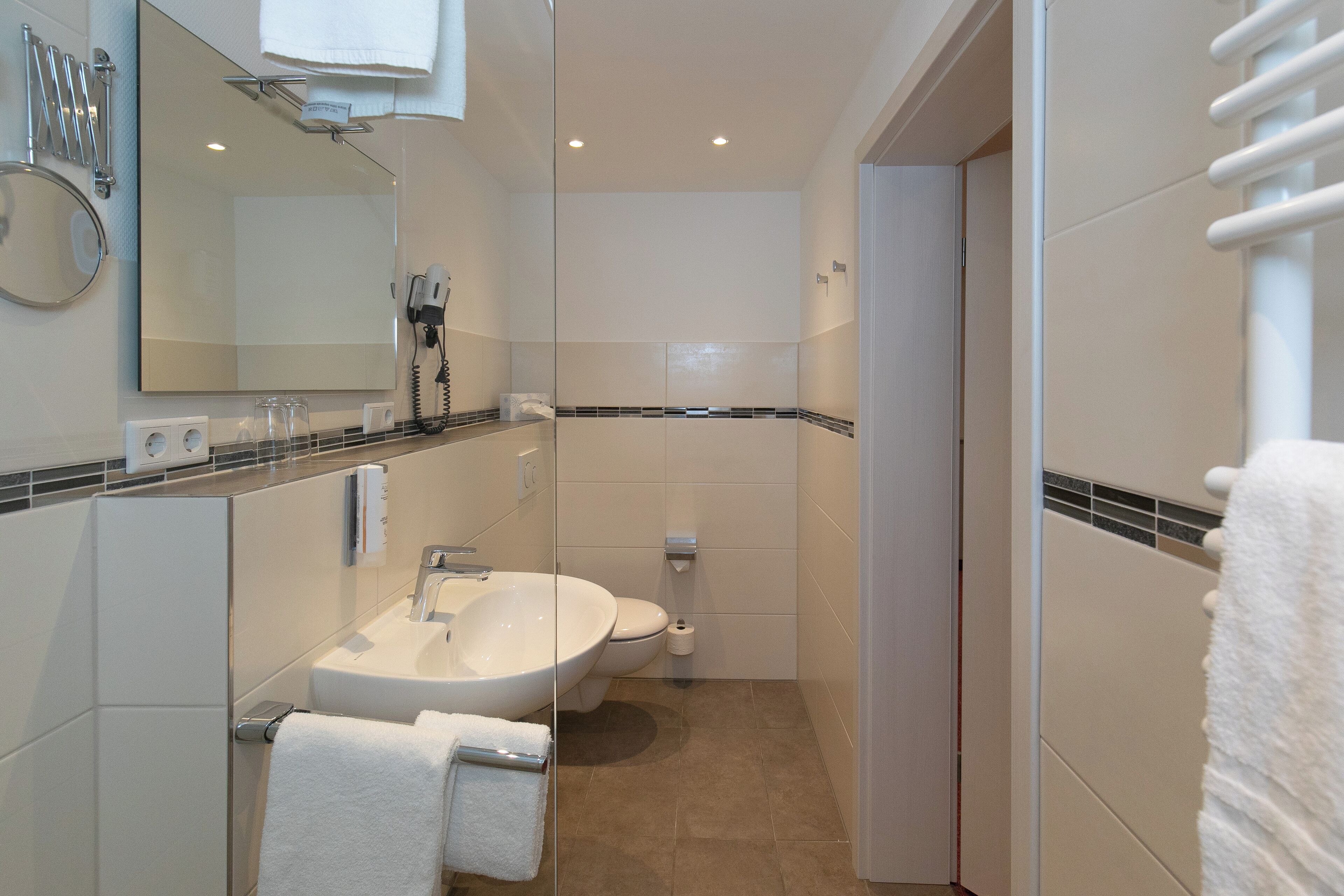 A modern bathroom with a large mirror, a sink, and a toilet. There are towels hanging on a rack and a towel dispenser.