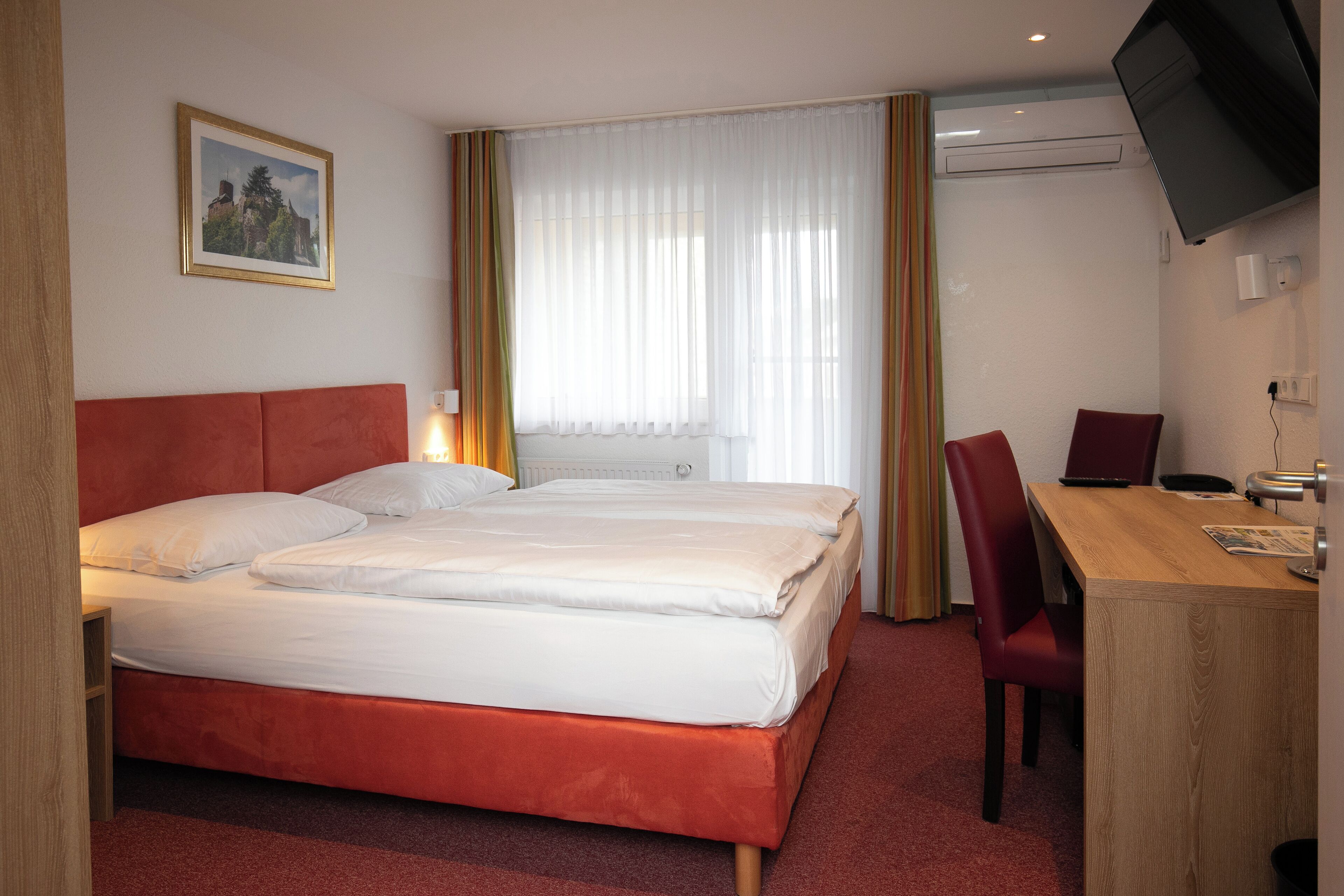 A hotel room with a double bed, a desk with a chair, a television mounted on the wall, and a window with curtains.