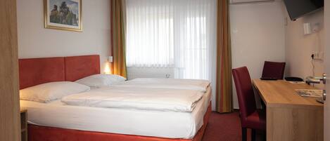 A hotel room with a double bed, a desk with a chair, a television mounted on the wall, and a window with curtains.