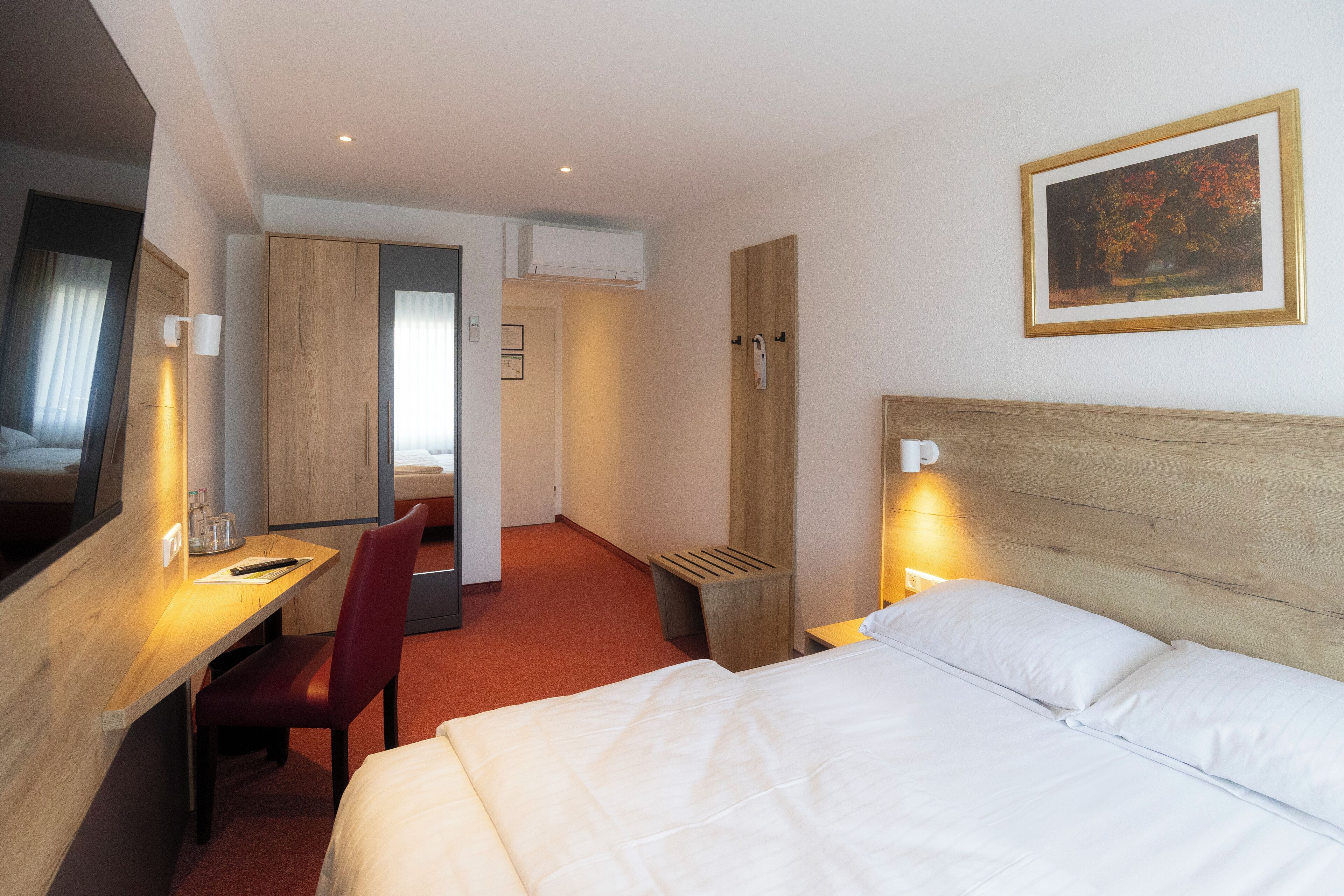 A hotel room with a bed, a desk, a chair, a TV, a painting on the wall, and an air conditioning unit.