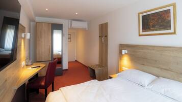 A hotel room with a bed, a desk, a chair, a TV, a painting on the wall, and an air conditioning unit.