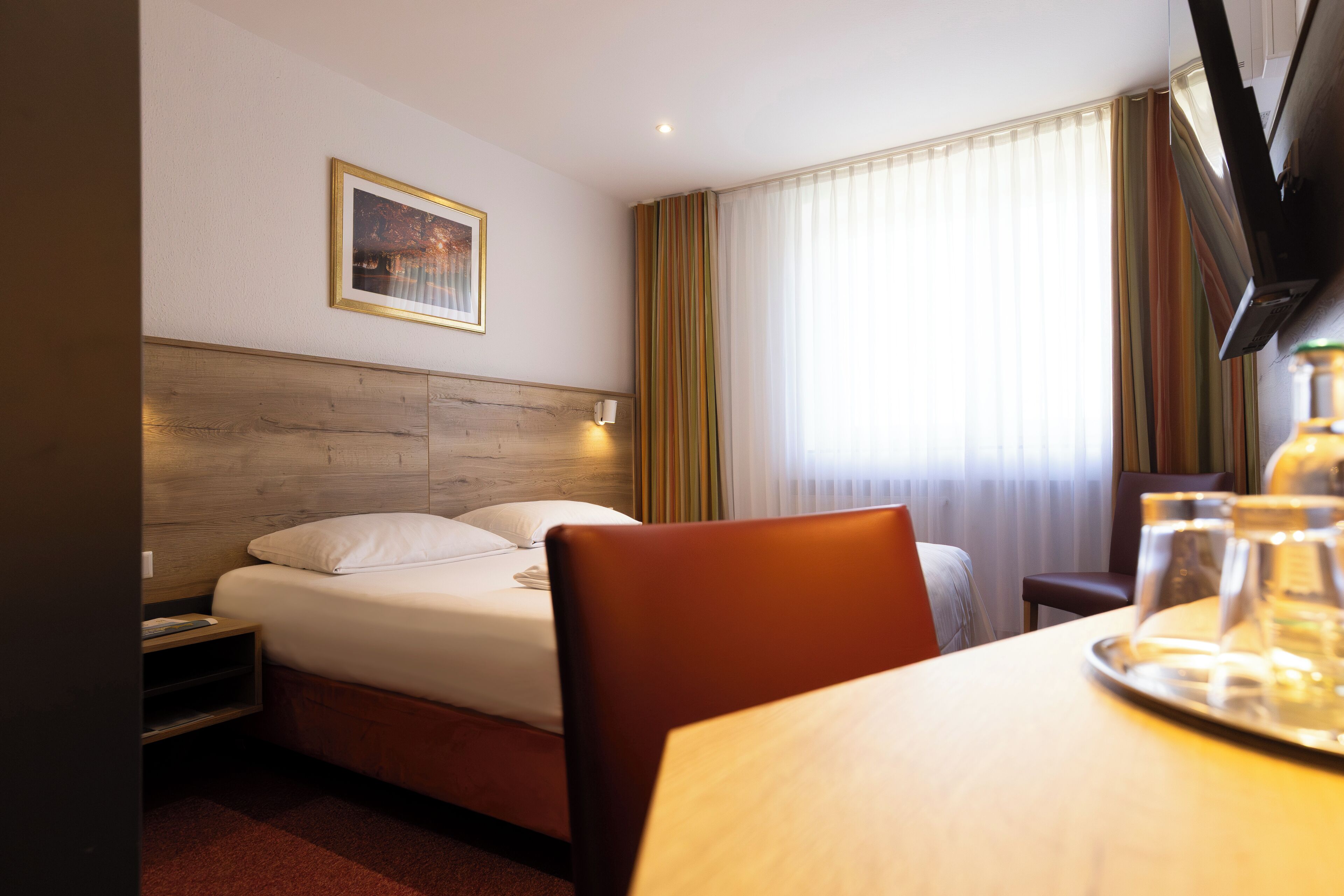 Een hotelkamer met een bed, een nachtkastje, een stoel, een eettafel met glazen en een schilderij aan de muur.