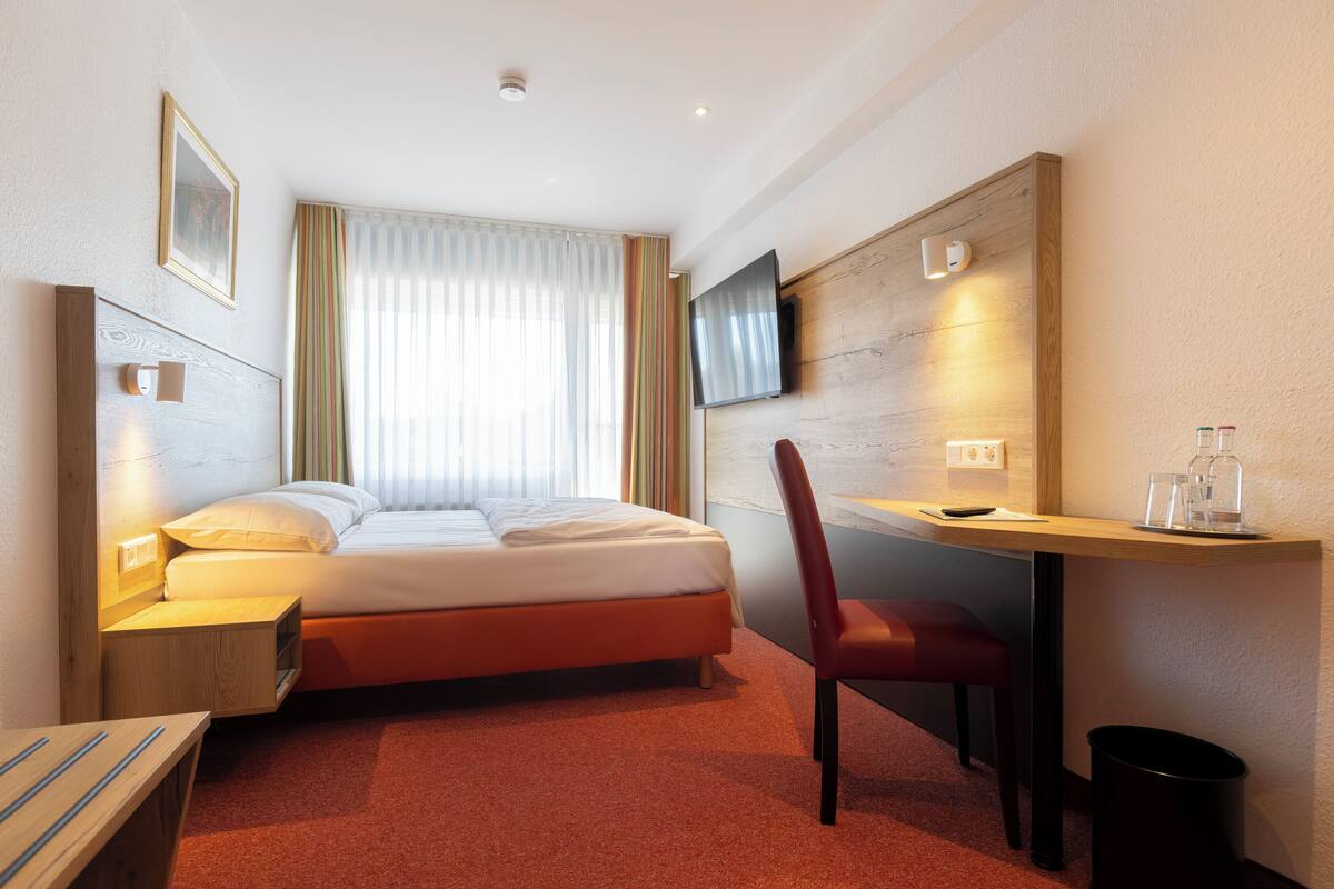 Habitación de hotel con cama, escritorio, silla, televisor montado en la pared y ventana con cortinas.