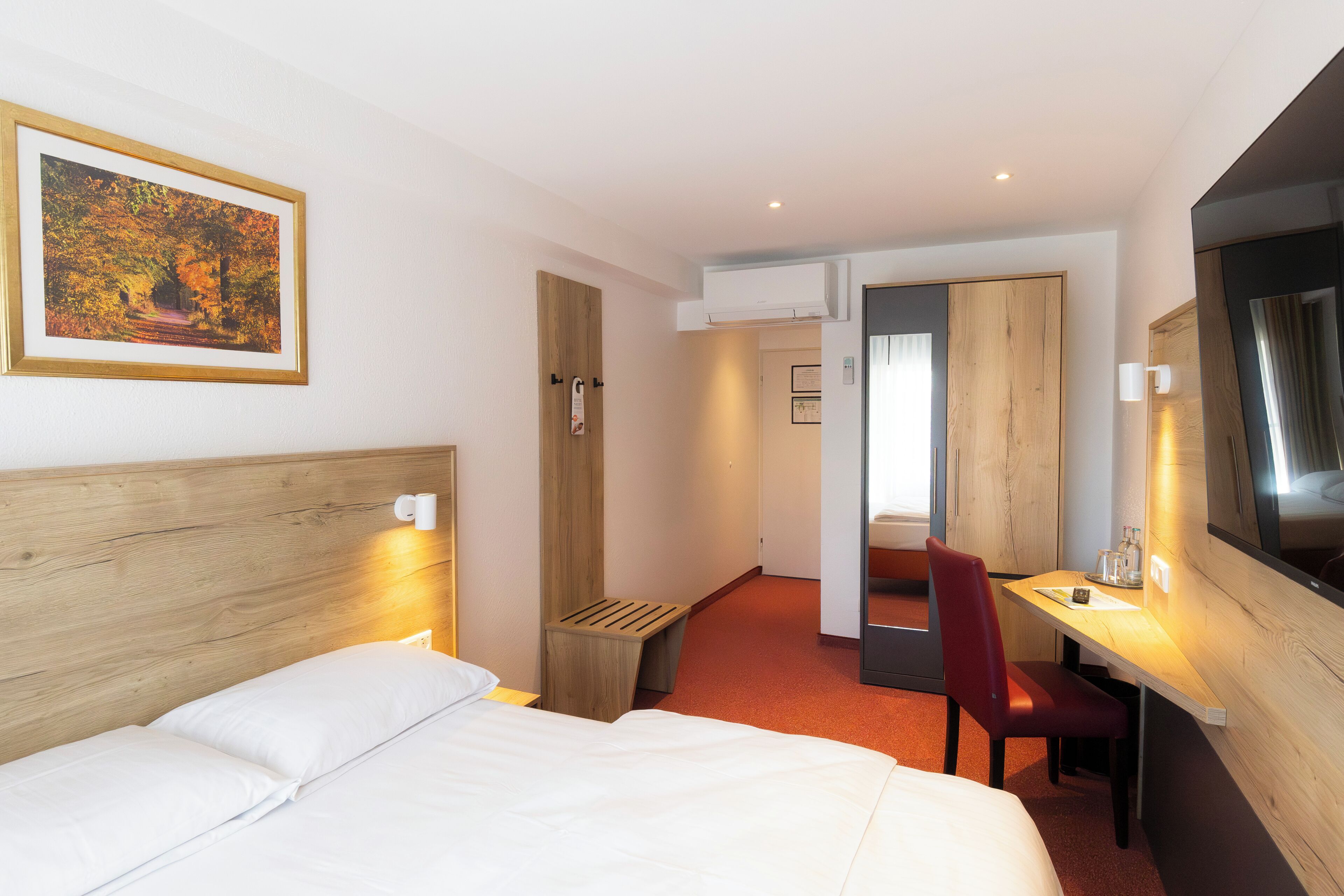 A hotel room with a wooden headboard, a bed, a desk with a chair, a TV, and a framed picture on the wall.