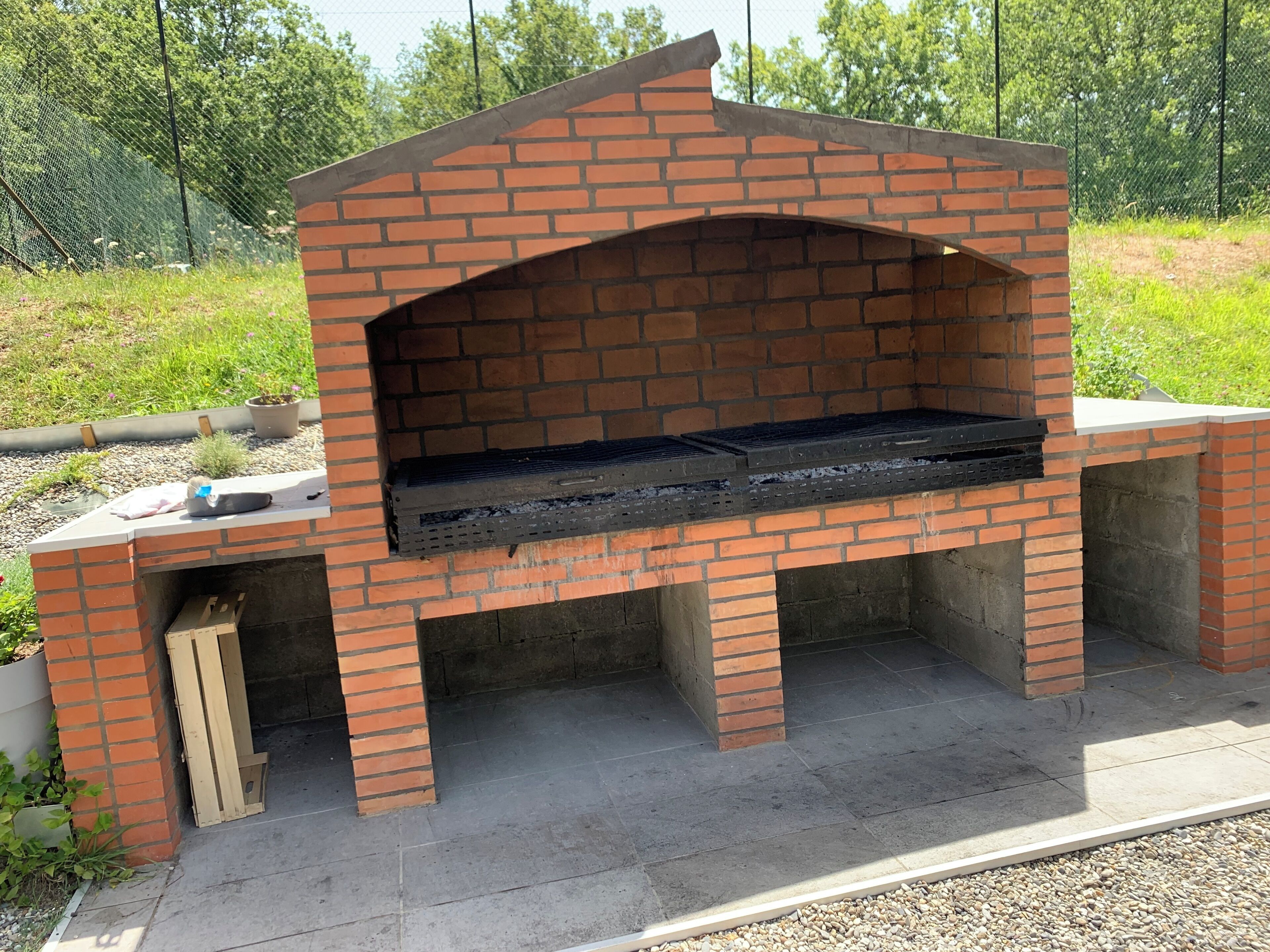 A brick-built outdoor oven with a flat top and a chimney, surrounded by gravel and greenery.