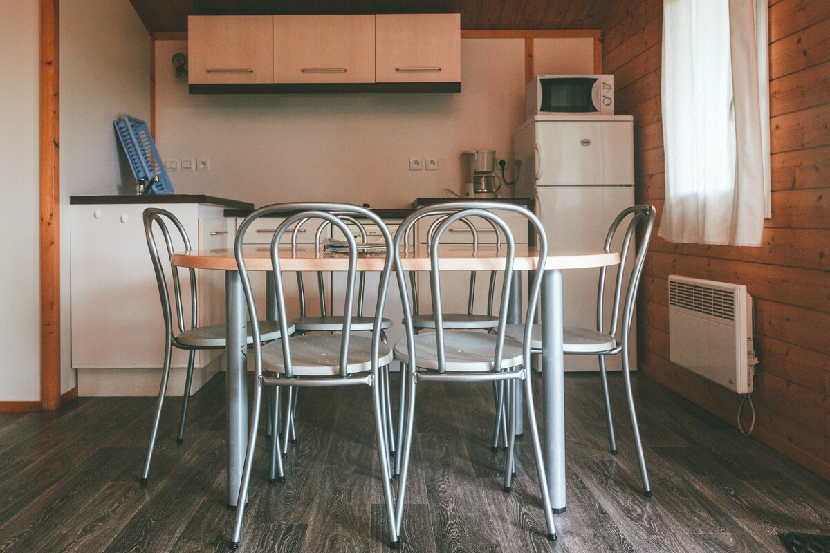 A compact kitchen with a wooden ceiling, a dining table with four chairs, a small refrigerator, and a coffee maker.