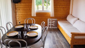 Interior de una cabaña de madera con una mesa de comedor para cuatro personas, un sofá y una ventana con cortinas.