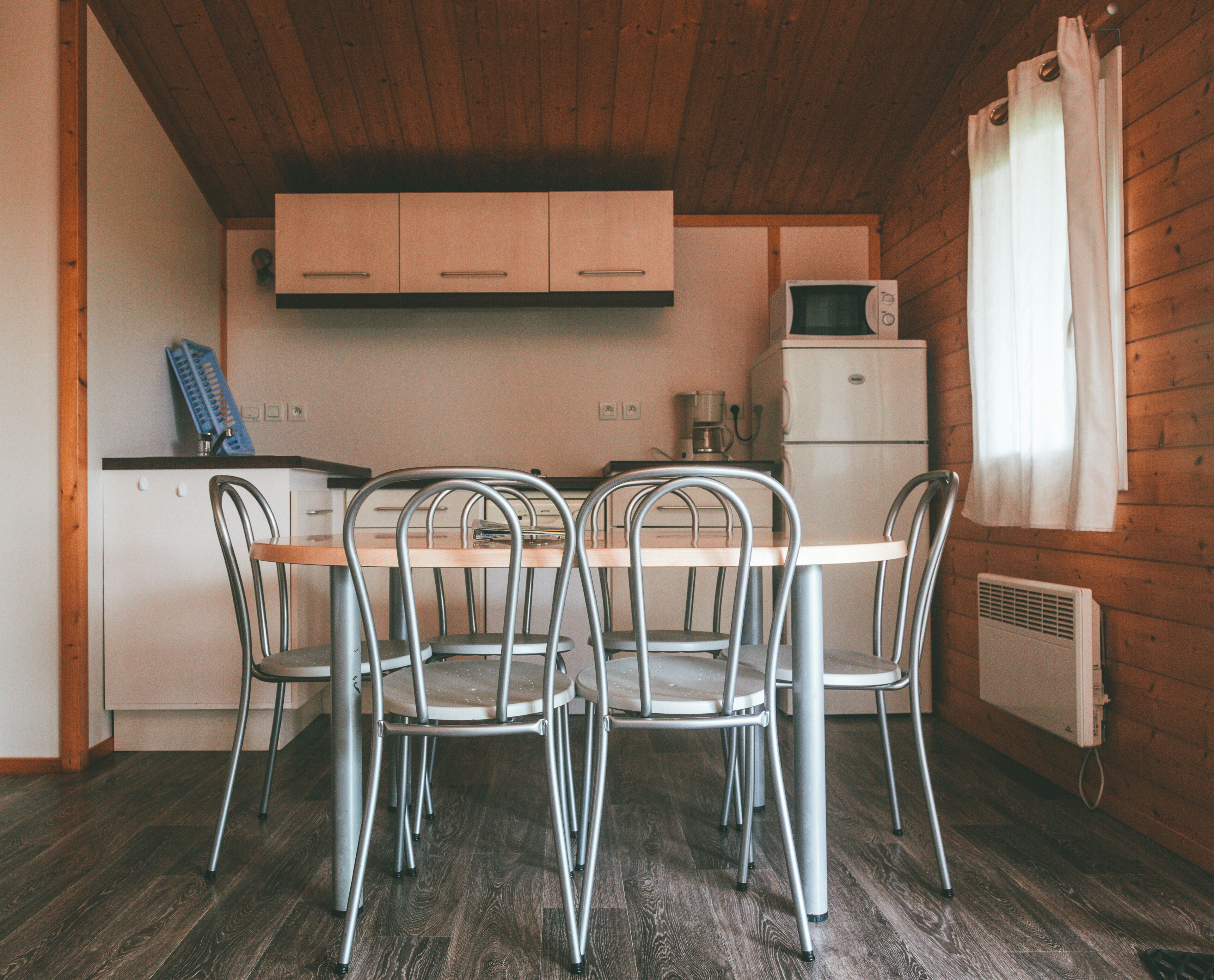 A compact kitchen with a wooden ceiling, a dining table with four chairs, a small refrigerator, and a coffee maker.