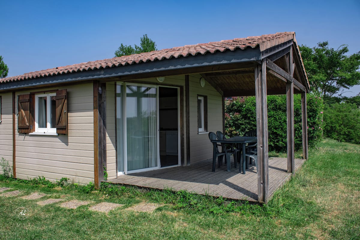 A single-story wooden house with a covered patio, a sliding glass door leading to a kitchen, and a small outdoor seating area.