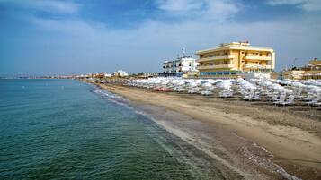 Un hotel a la vora de la platja, amb diverses plantes, una platja sorrenca i files de gandules.