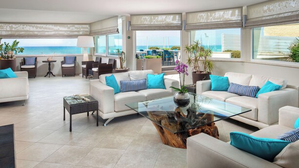 A spacious living area with a glass coffee table, white sofas with blue cushions, and a view of the ocean. - Hotel Miramare (Latina)