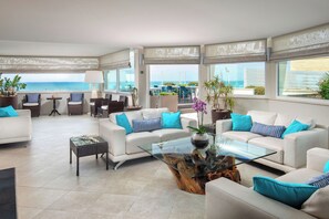 A spacious living area with a glass coffee table, white sofas with blue cushions, and a view of the ocean. - Hotel Miramare (Latina)
