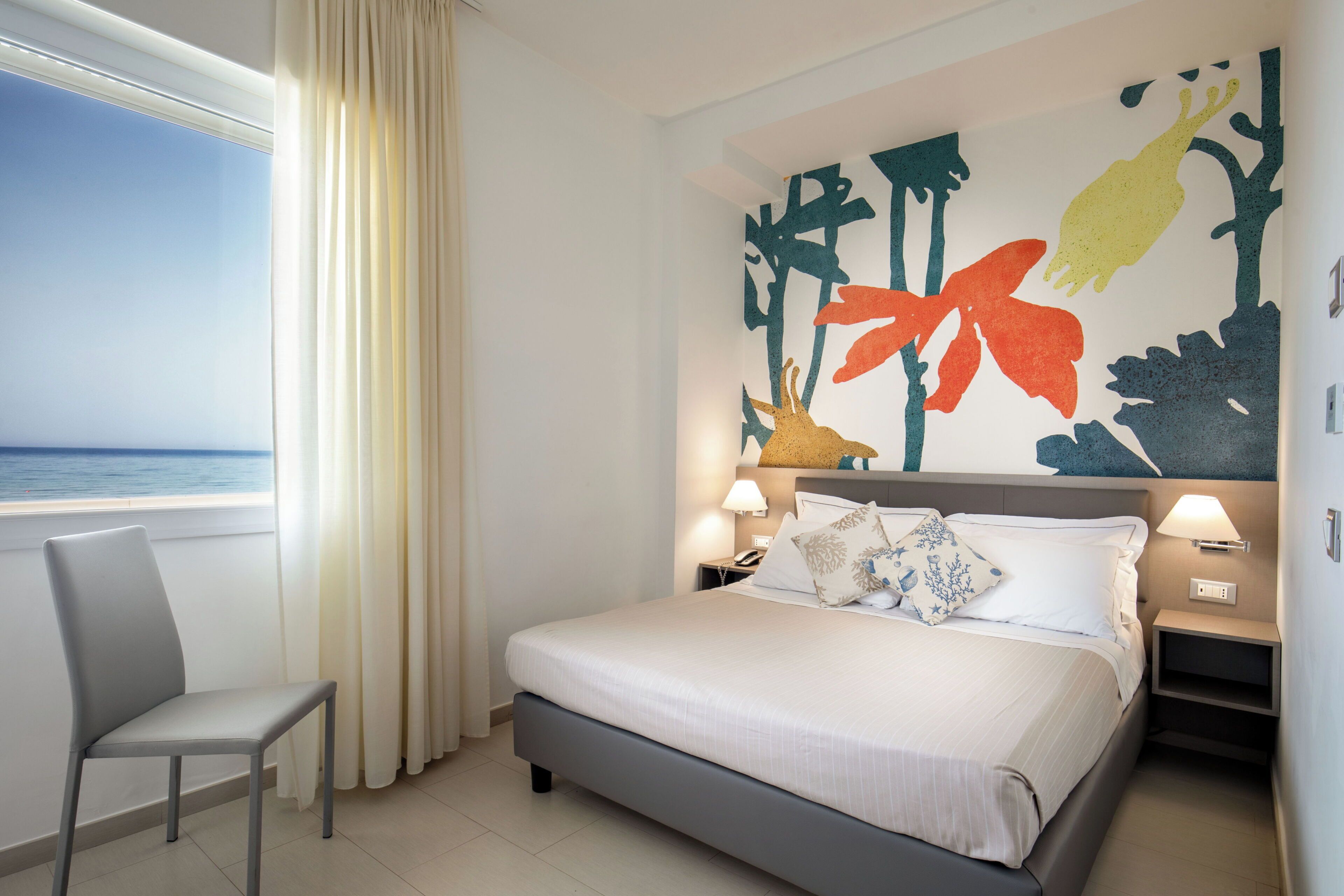 Habitación de hotel con cama, una silla, mesita de noche, pared con un mural colorido y ventana con cortinas.