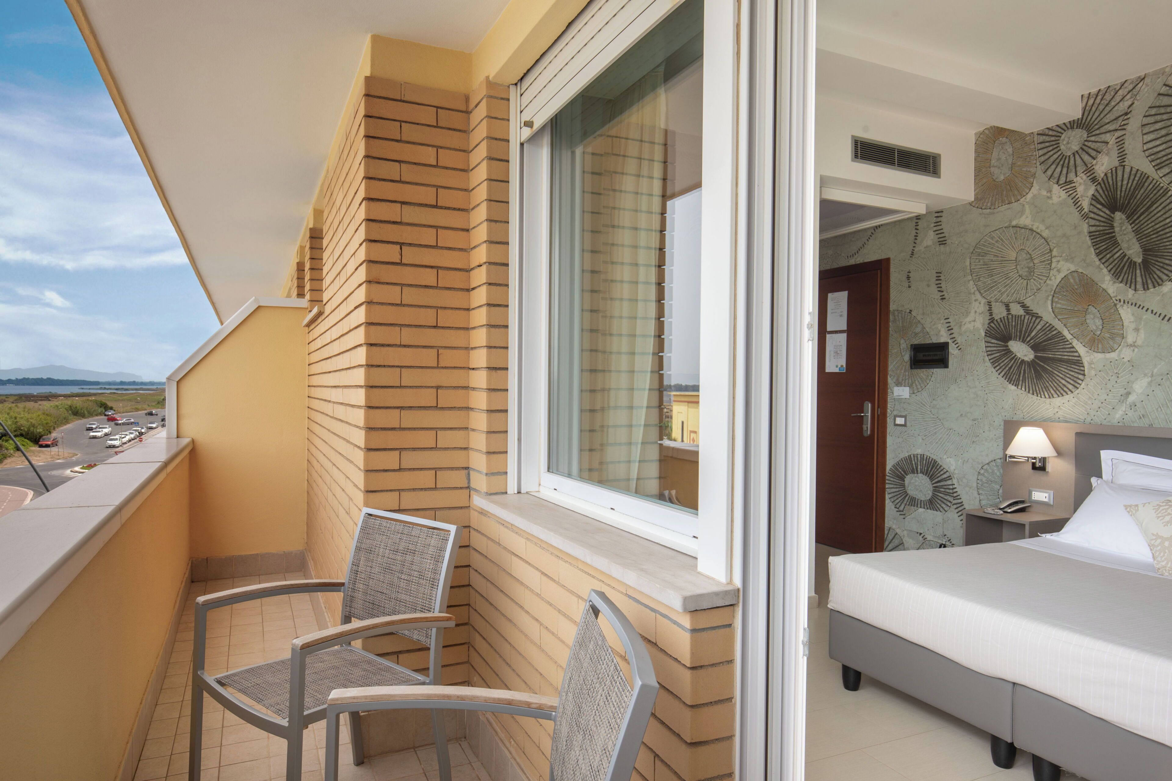 A balcony with a view of a road and a bed with a headboard and a nightstand with a lamp.