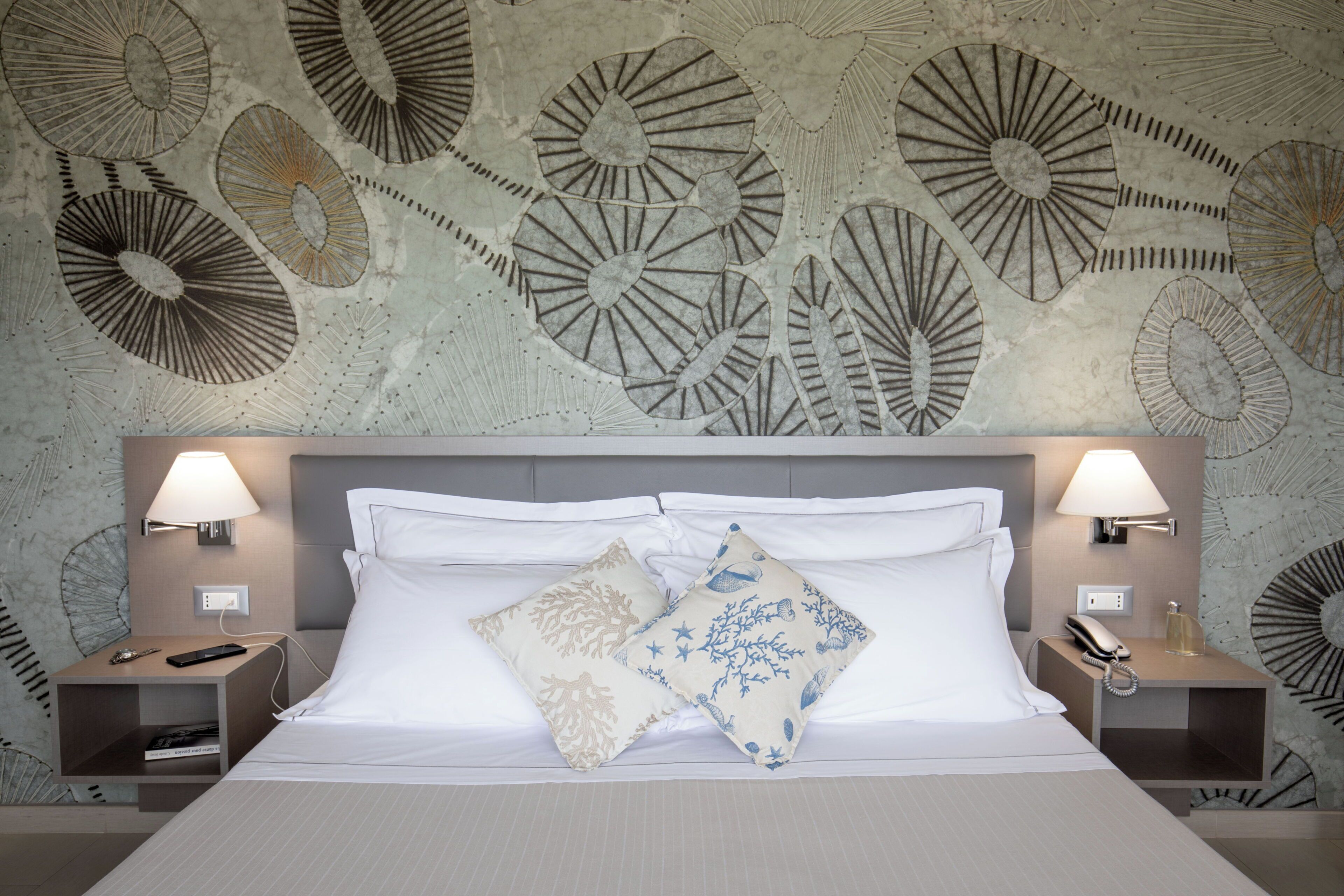 A hotel room with a large bed, two bedside tables, a wall with a decorative pattern, and two wall-mounted lamps.