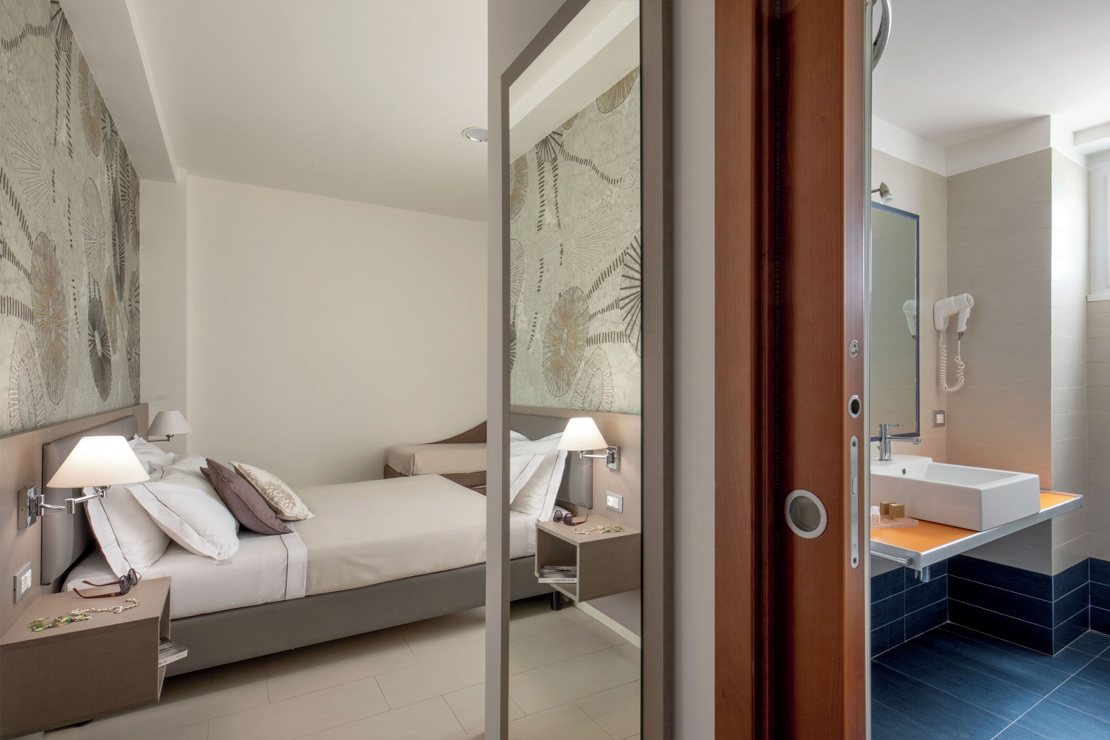 Een hotelkamer met een bed, nachtkastje, lamp en een badkamer met wastafel en spiegel.