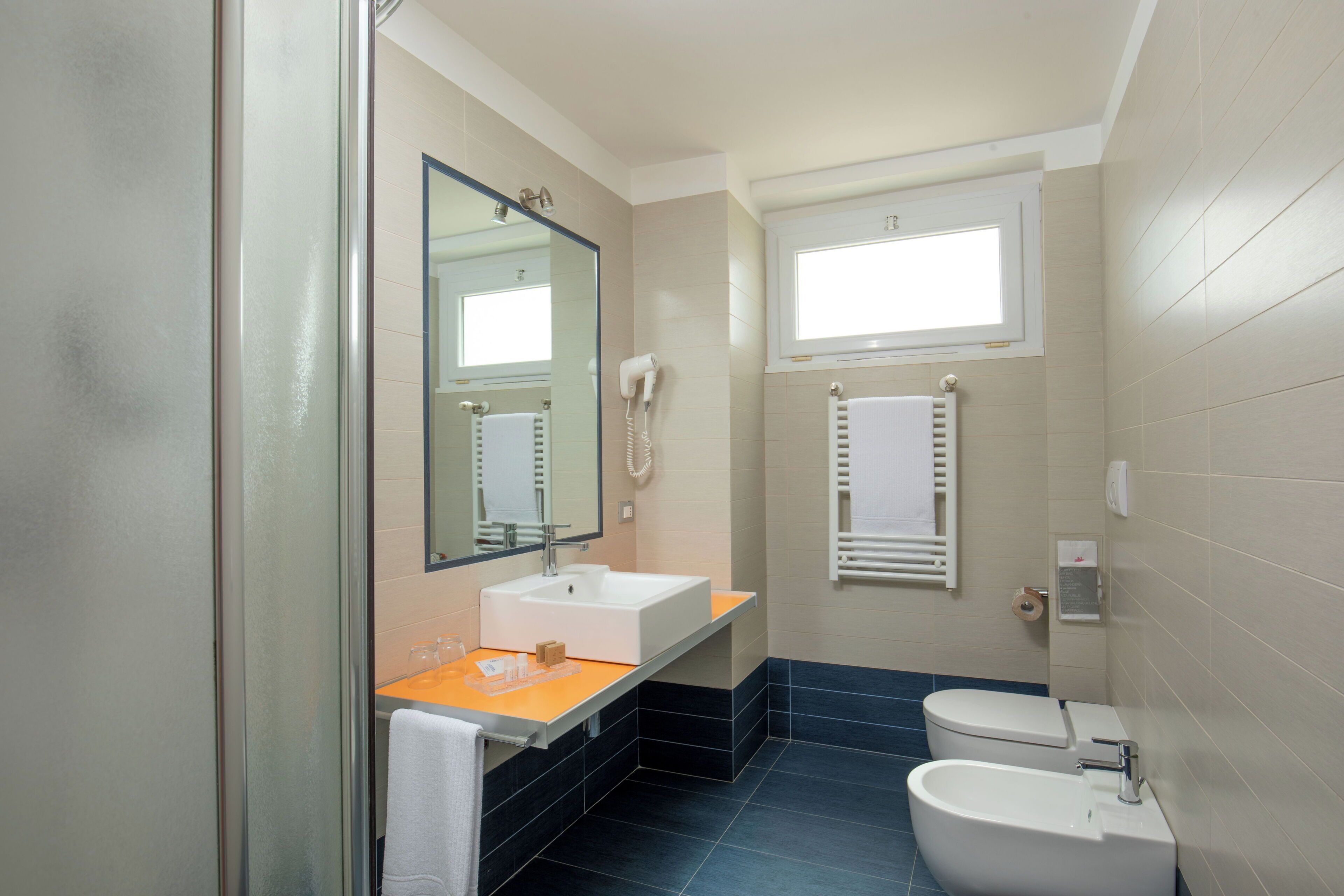 Un baño con cabina de ducha de vidrio, un lavabo blanco, un inodoro y una ventana sobre el inodoro.